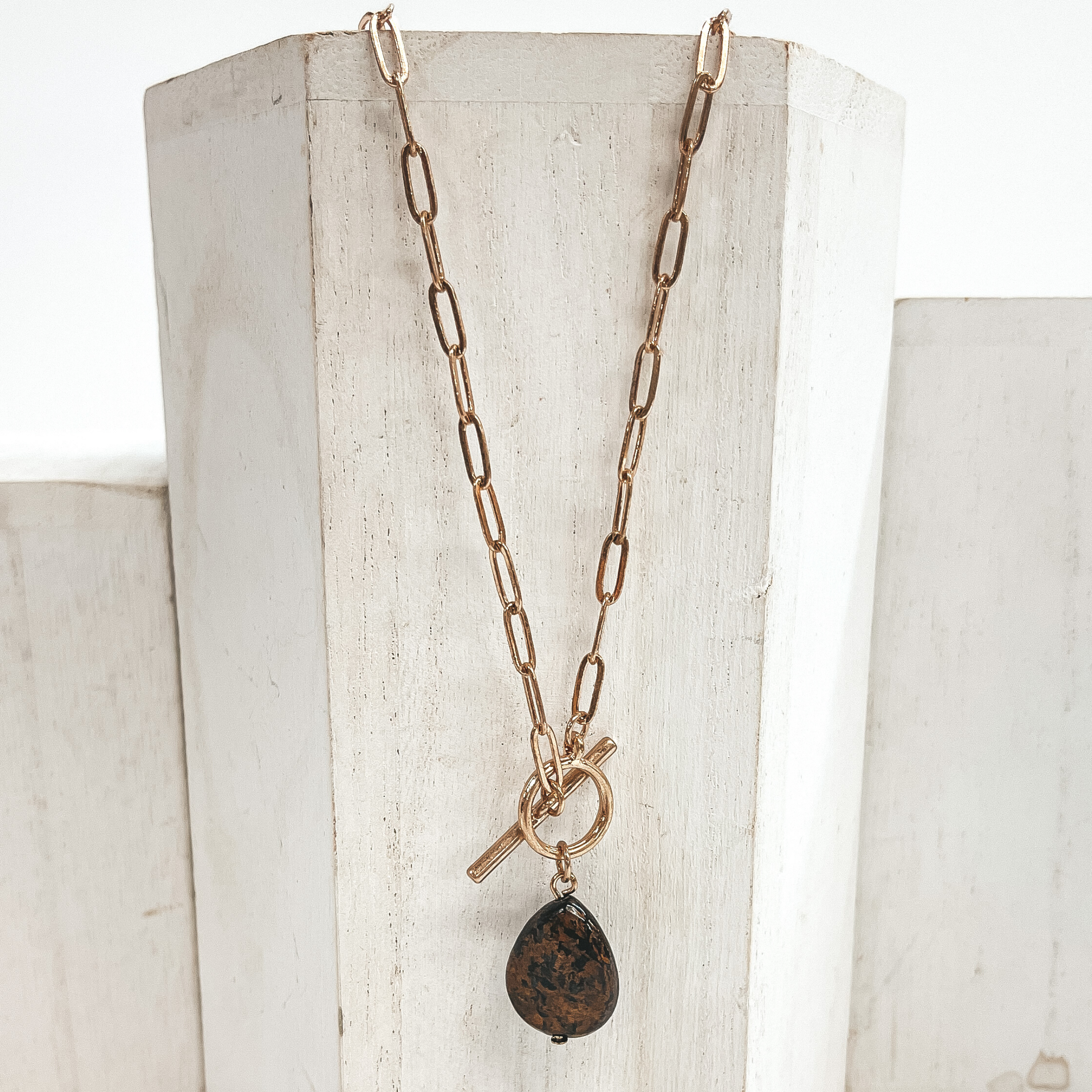 Gold paperclip chain necklace with toggle clasp and  natural stone teardrop pendant in brown. Taken on  a white block and white background.