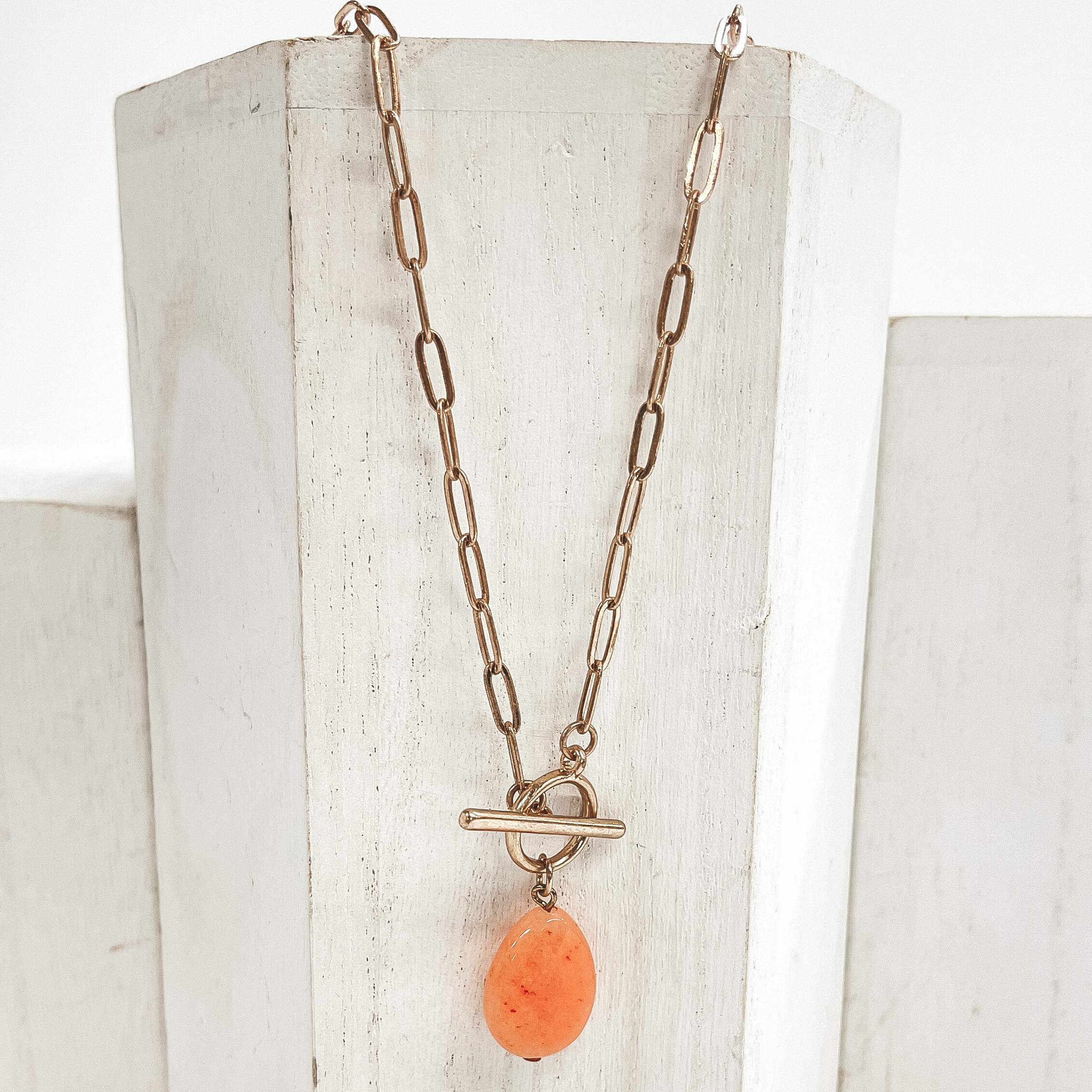 Gold paperclip chain necklace with toggle clasp and  natural stone teardrop pendant in coral orange.  Taken on  a white block and white background.