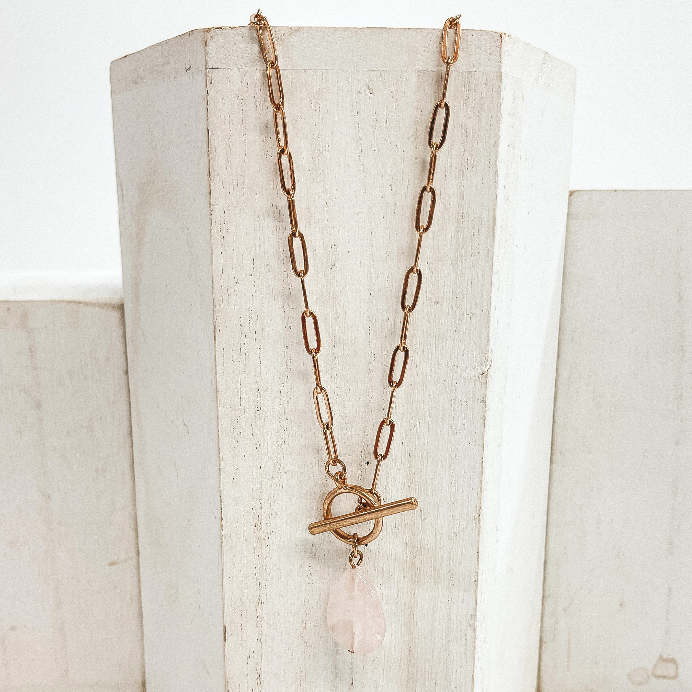 Gold paperclip chain necklace with toggle clasp and  natural stone teardrop pendant in light pink.  Taken on  a white block and white background.