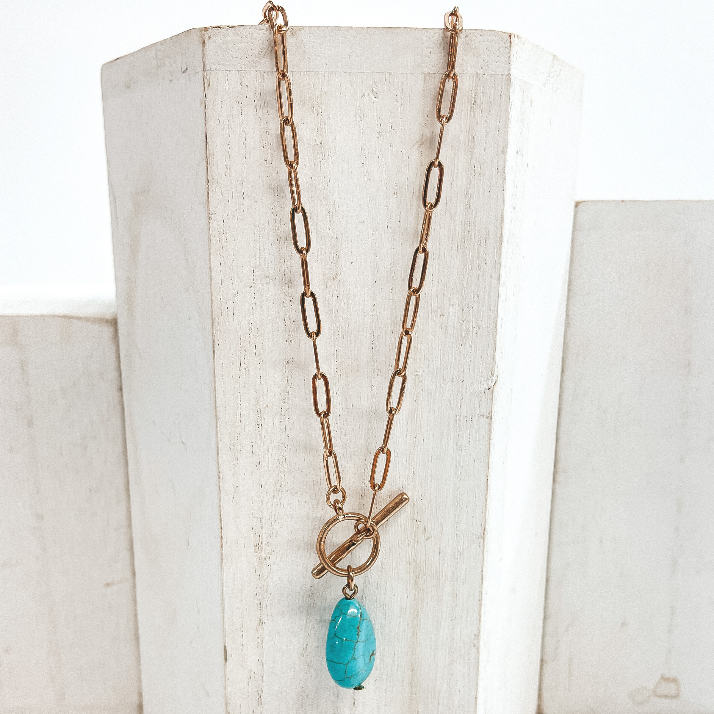 Gold paperclip chain necklace with toggle clasp and  natural stone teardrop pendant in turquoise.  Taken on  a white block and white background.