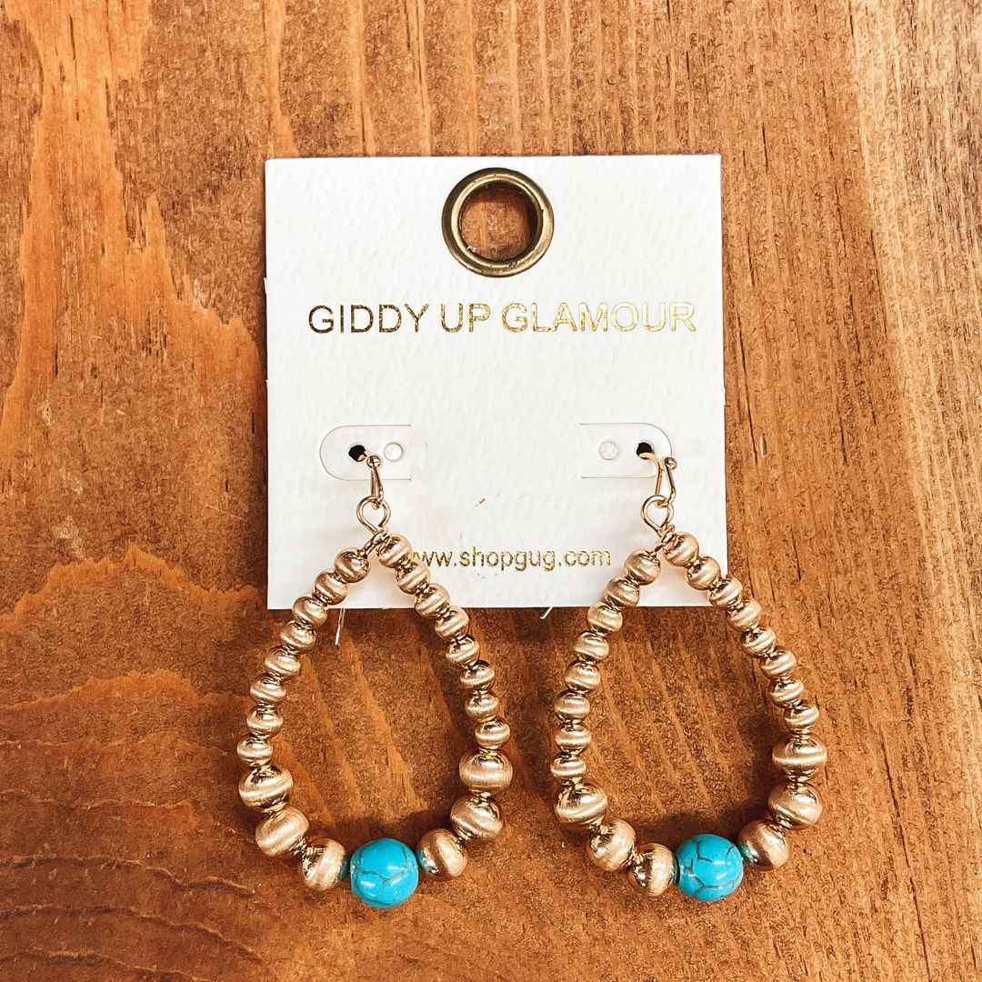 Gold beaded teardrop earrings with fish hook backing,  there is one faux turquoise bead in the center.  Taken on top of a brown block.