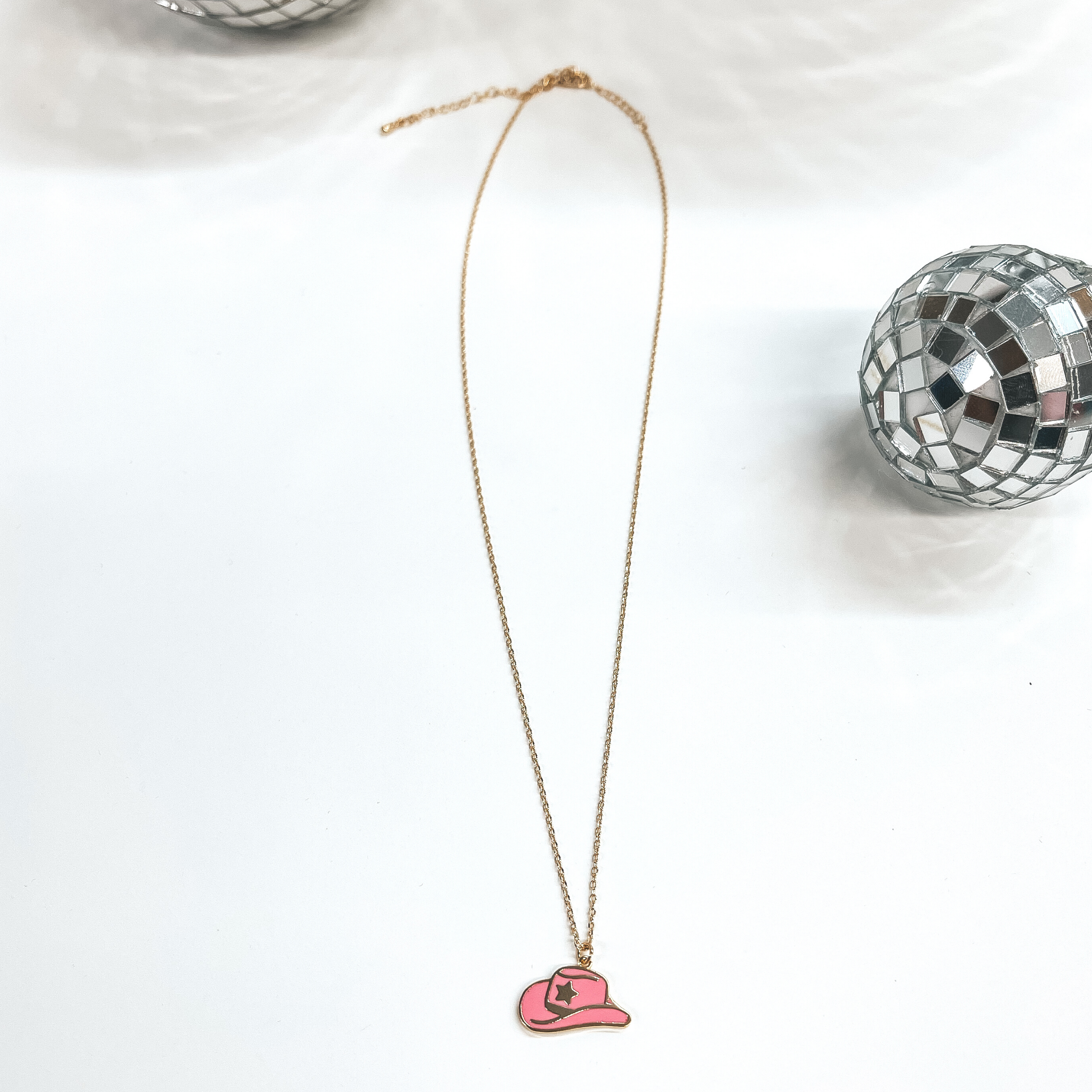 Small gold chain necklace with a pink hat pendant,  the hat pendant has a gold star in the center.  Taken on a white background with disco balls as decor.