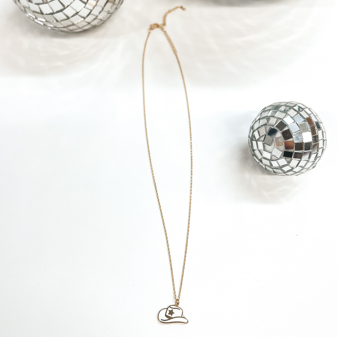 Small gold chain necklace with a white hat pendant,  the hat pendant has a gold star in the center.  Taken on a white background with disco balls as decor.