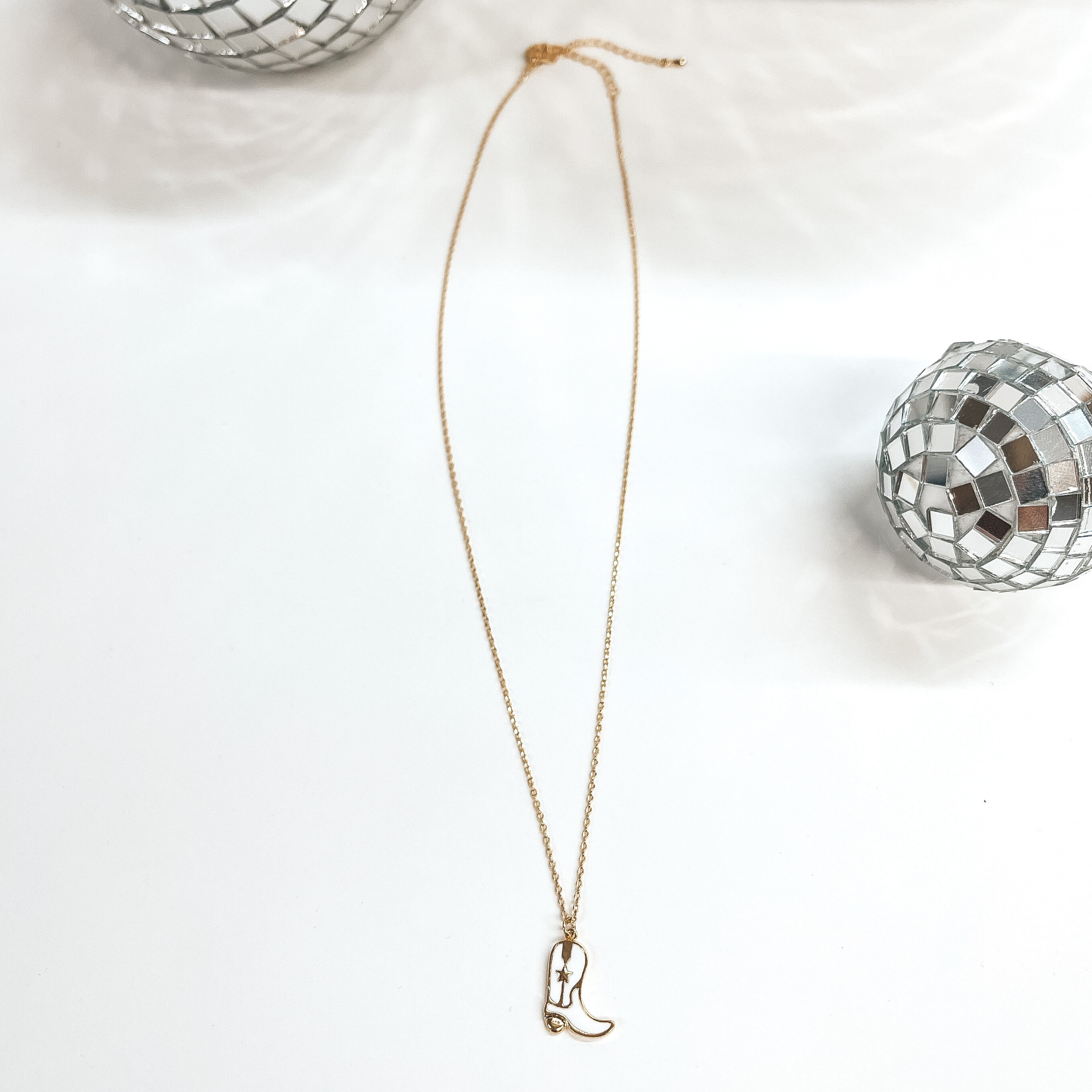 Small gold chain necklace with a white boot pendant,  the boot pendant has a gold star in the center.  Taken on a white background with disco balls as decor.