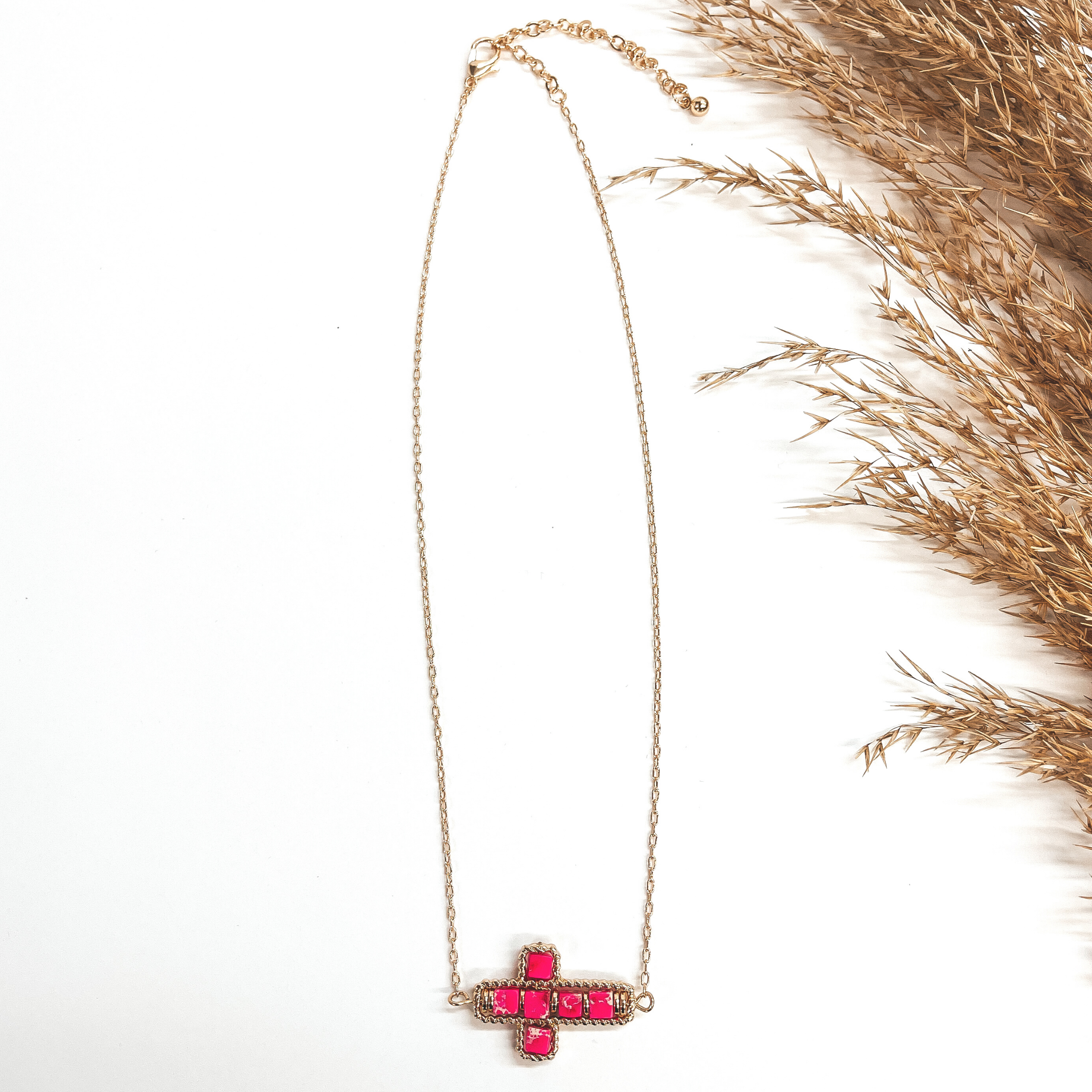 This a gold chain necklace with a cross pendant in  the center. The cross pendant has a gold rope  texture around and has semi- precious stones in  fuchsia. It is taken on a white background and a  brown plant in the side as decor.