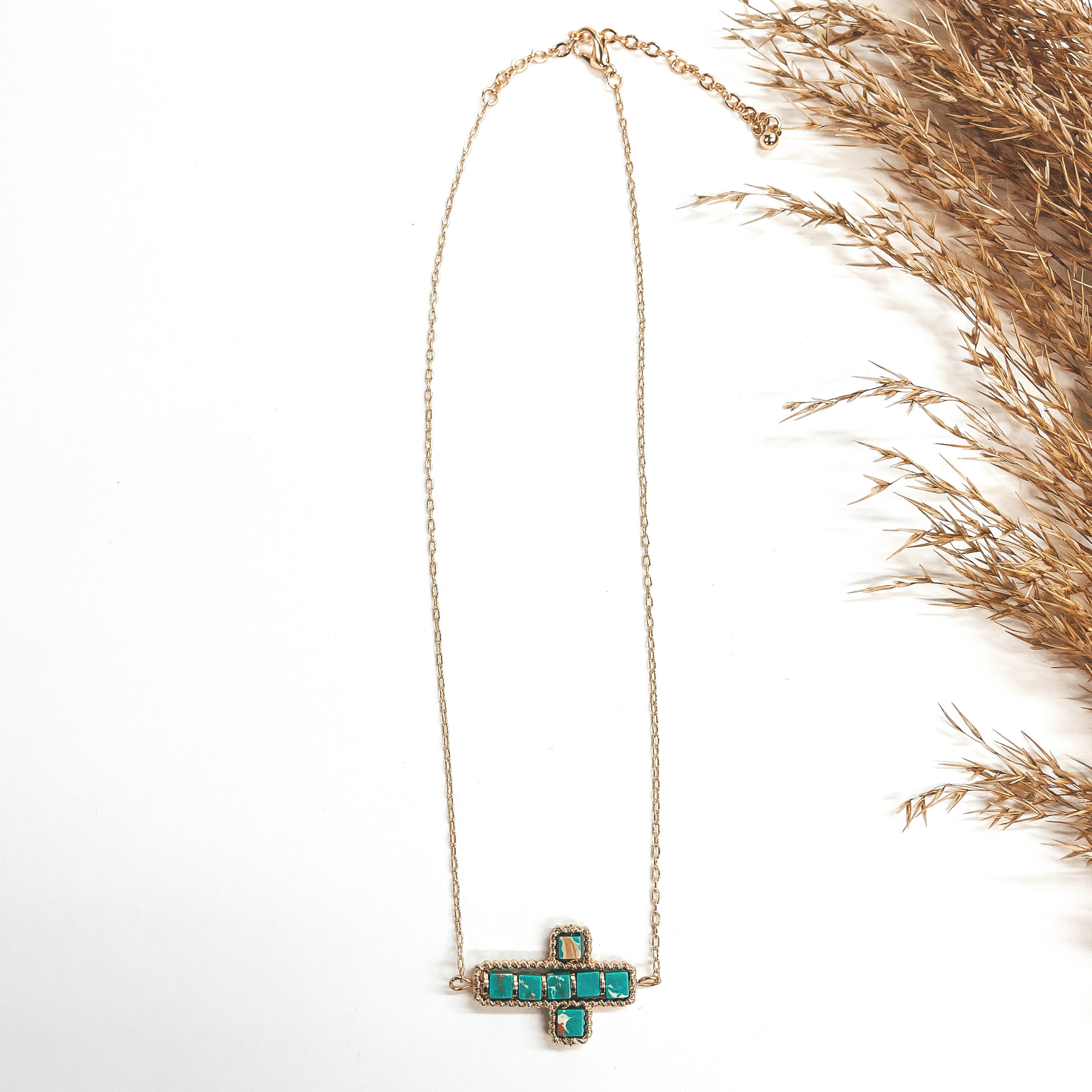 This a gold chain necklace with a cross pendant in  the center. The cross pendant has a gold rope  texture around and has semi- precious stones in  turquoise. It is taken on a white background and a  brown plant in the side as decor.
