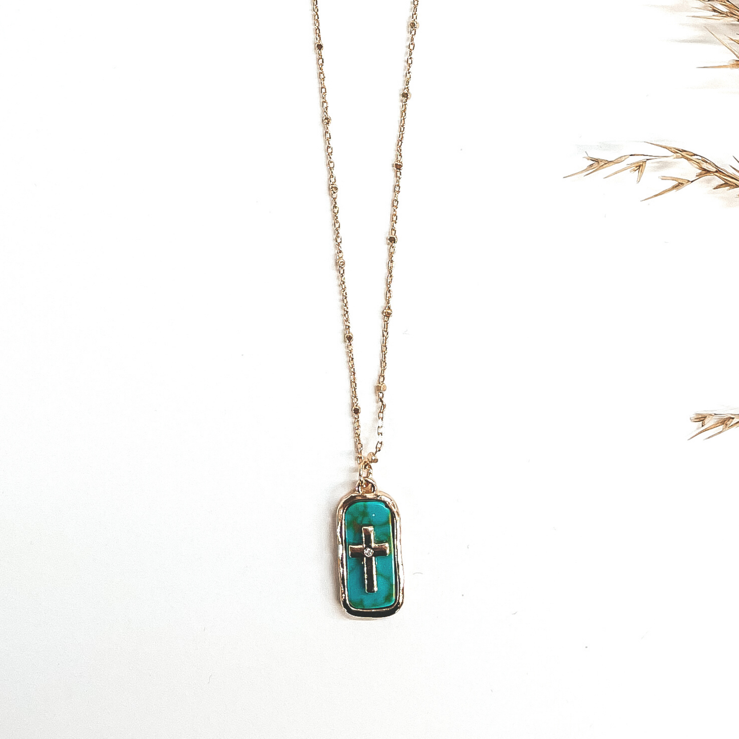This a gold chain necklace with a rectangular pendant, pendant is a semi-precious stone in turquoise. It  has a gold cross in the center with a small clear  crystal in the center of the cross. The chain has gold spacers. This is  taken on a white background with a brown plant in the  side as decor.