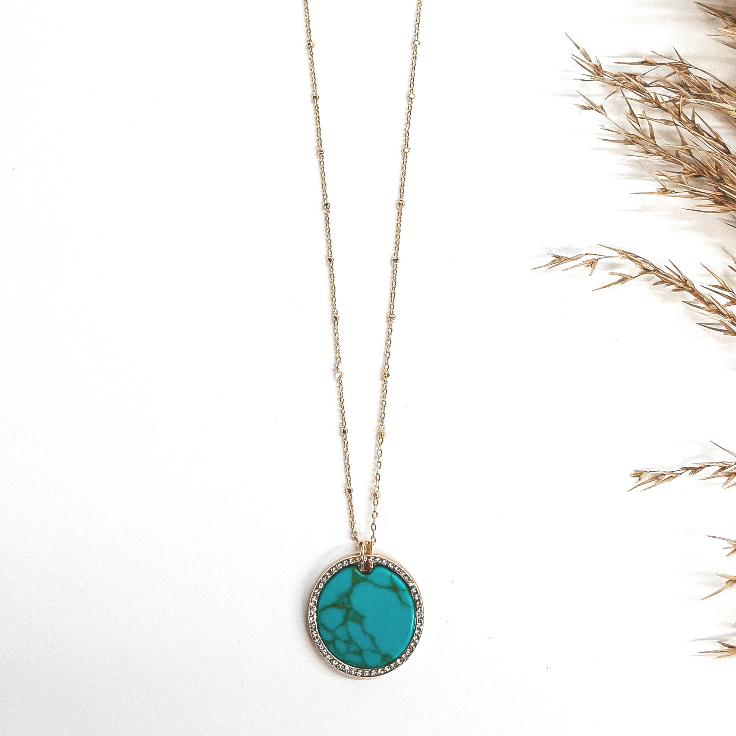 This a gold chain necklace with a circle pendant, the  circle pendant is a semi-precious stone in turquoise.  The stone pendant is in a gold settings and has CZ  crystals around. The chain has gold spacers. This is  taken on a white background with a brown plant in the  side as decor.