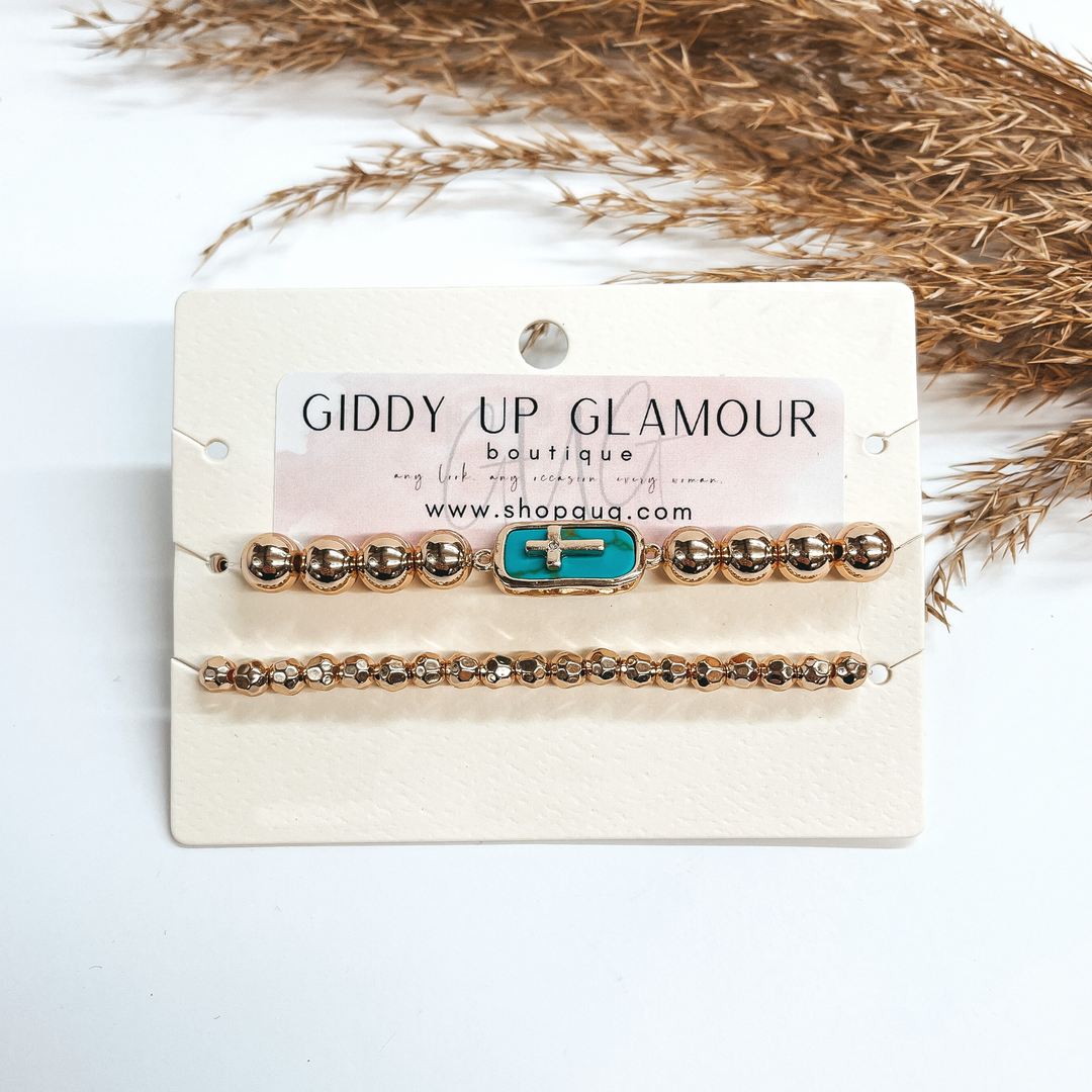 This a gold beaded bracelet set of two, the bottom  bracelet has smaller gold shiny beads. The top  bracelet has big gold beads with a rectangular pendant. The rectangular pendant is in a gold setting and semi-  precious turquoise stone. The pendant has a  small gold cross in  the center and a small clear crystal in the center  as well. Taken on a white background with a brown  plant in the back as decor.