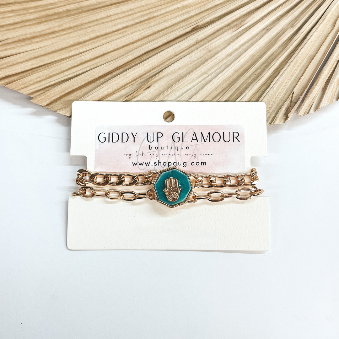 This a two gold chained magnetic bracelet, it comes  together with a semi-precious teal green stone in an  octagon shape. The octagon has a gold hand with an  eye in the center and a small clear crystal in the  center as well. Taken on a white background and  leaned up against a dried up palm leaf.