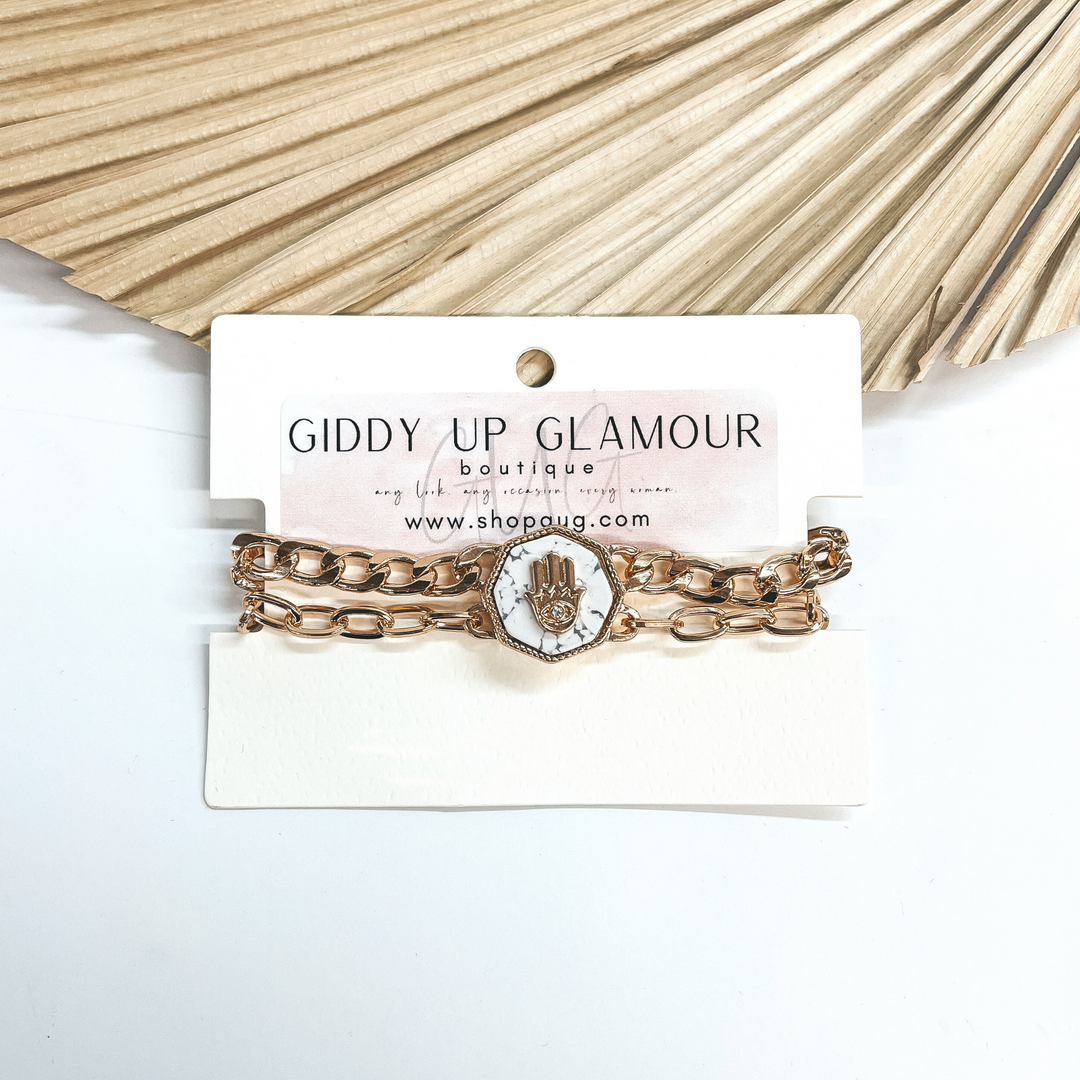 This a two gold chained magnetic bracelet, it comes  together with a semi-precious white stone in an  octagon shape. The octagon has a gold hand with an  eye in the center and a small clear crystal in the  center as well. Taken on a white background and  leaned up against a dried up palm leaf.