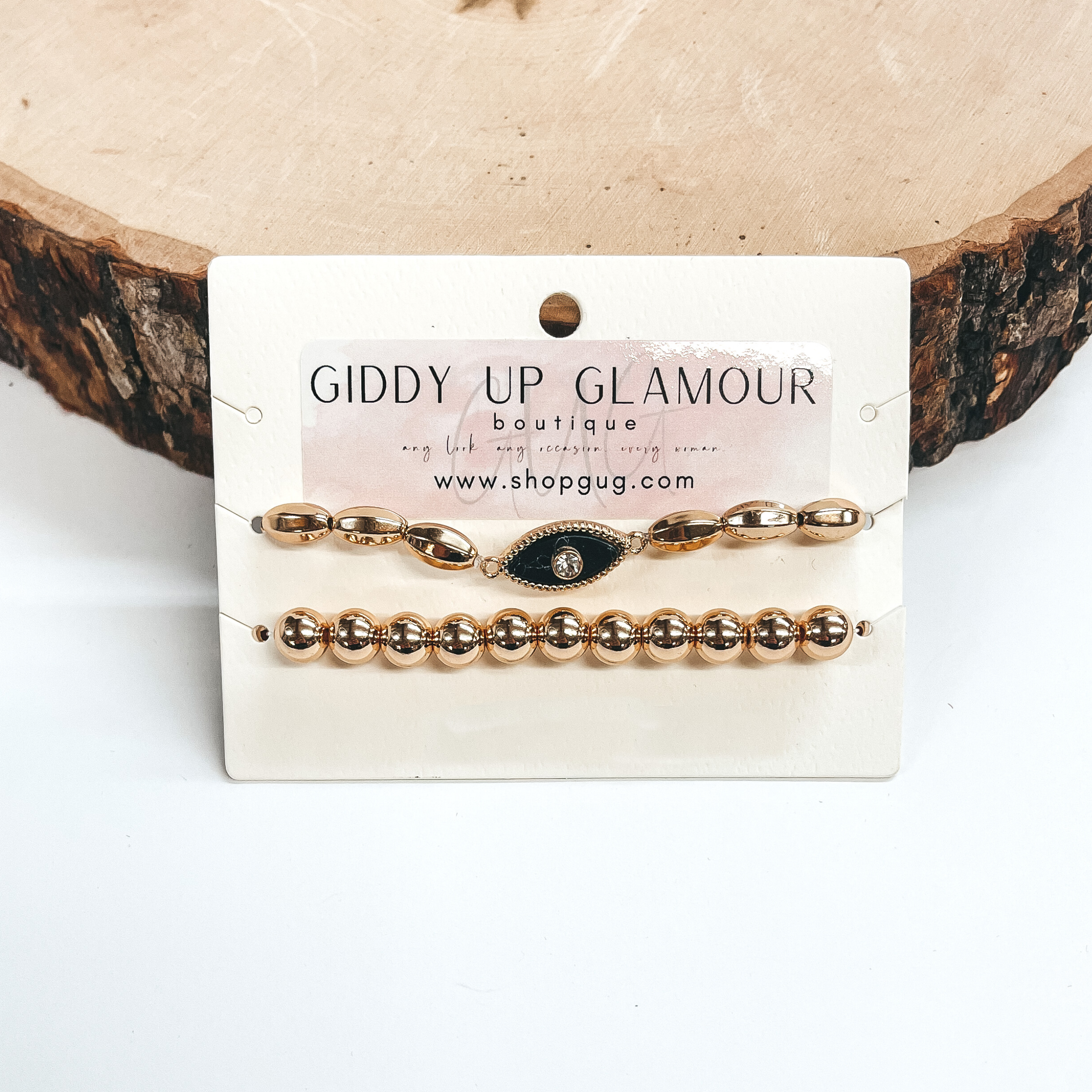 This a set of two bracelets with gold beads and an  evil eye pendant. The bottom bracelet are big gold  beads and the top bracelet has oval gold beads.  The evil eye is a semi-precious stone in black with  a CZ crystal in the middle. Taken on a white  background and leand up against a piece of wood.