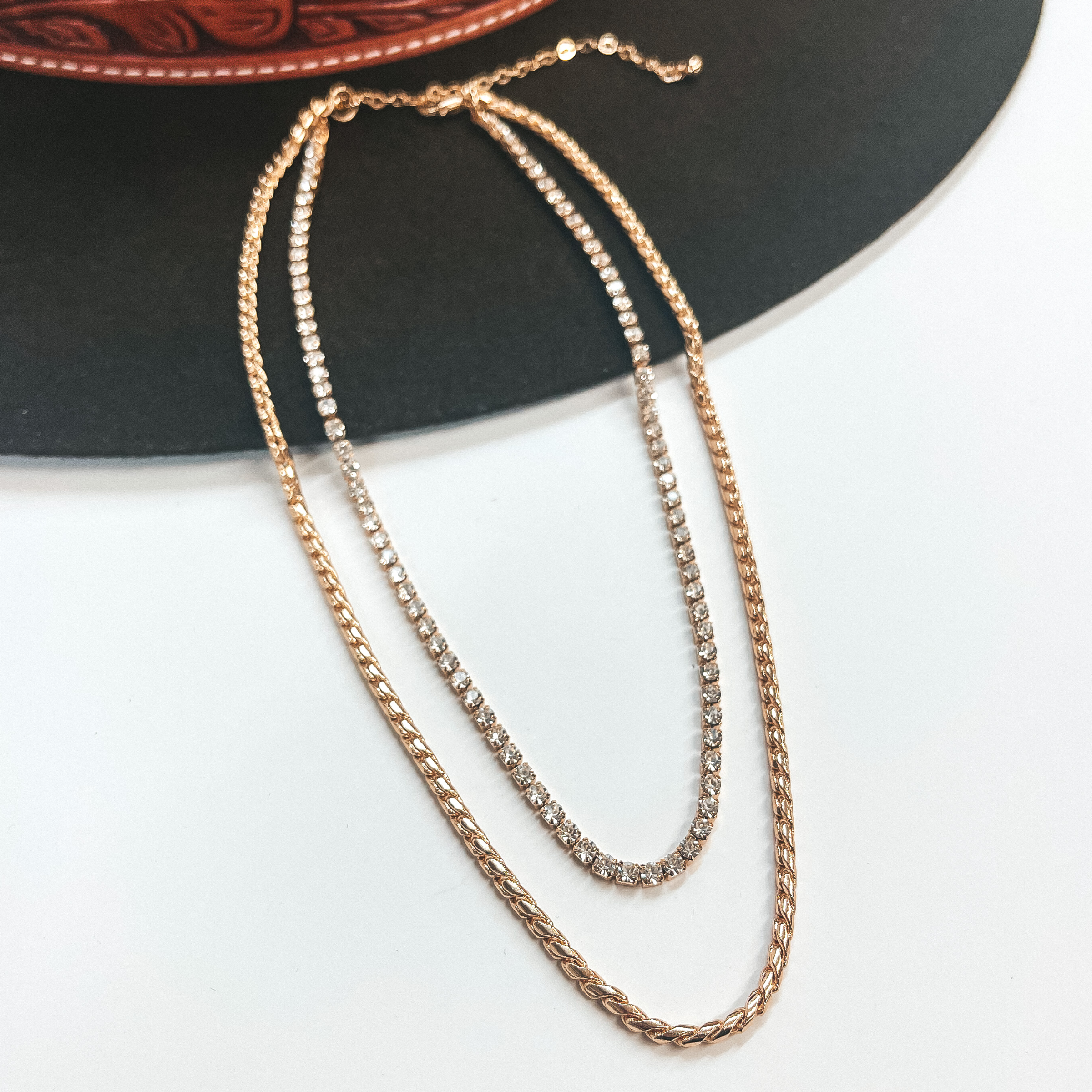 This is double layered gold chain necklace, the longer  chain is a braid textured chain. The smallest chain  has rhinestones all around. This necklace is pictured  laying on a dark brown hat and white background.