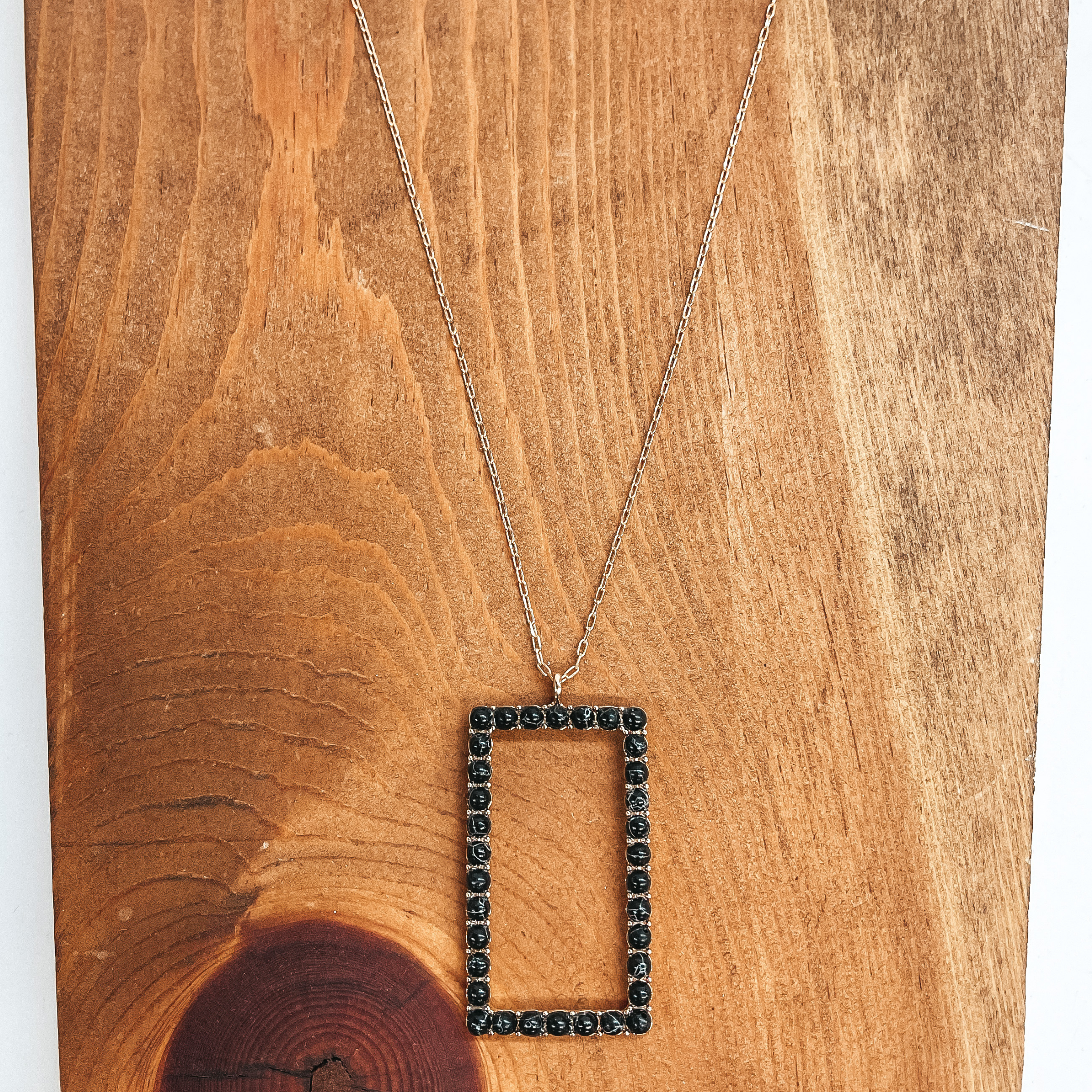 This is a long, thin, gold chain necklace with an  open rectangle pendant. The rectangle pendant has  semi-precious stones in black all around. This  necklace is taken on a brown block and white  background.