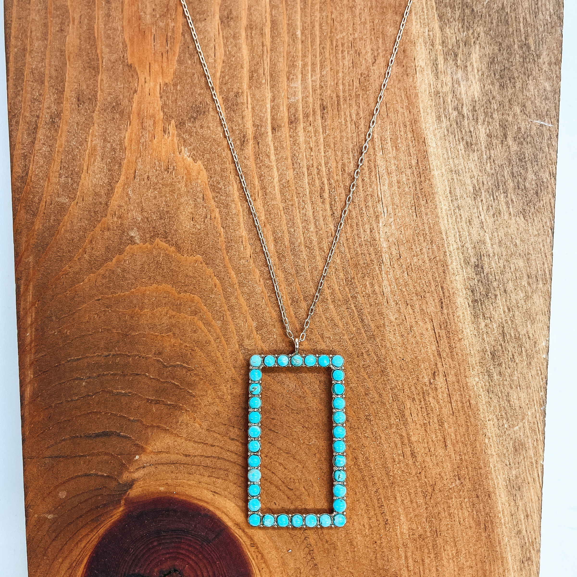 This is a long, thin, gold chain necklace with an  open rectangle pendant. The rectangle pendant has  semi-precious stones in turquoise all around. This  necklace is taken on a brown block and white  background.