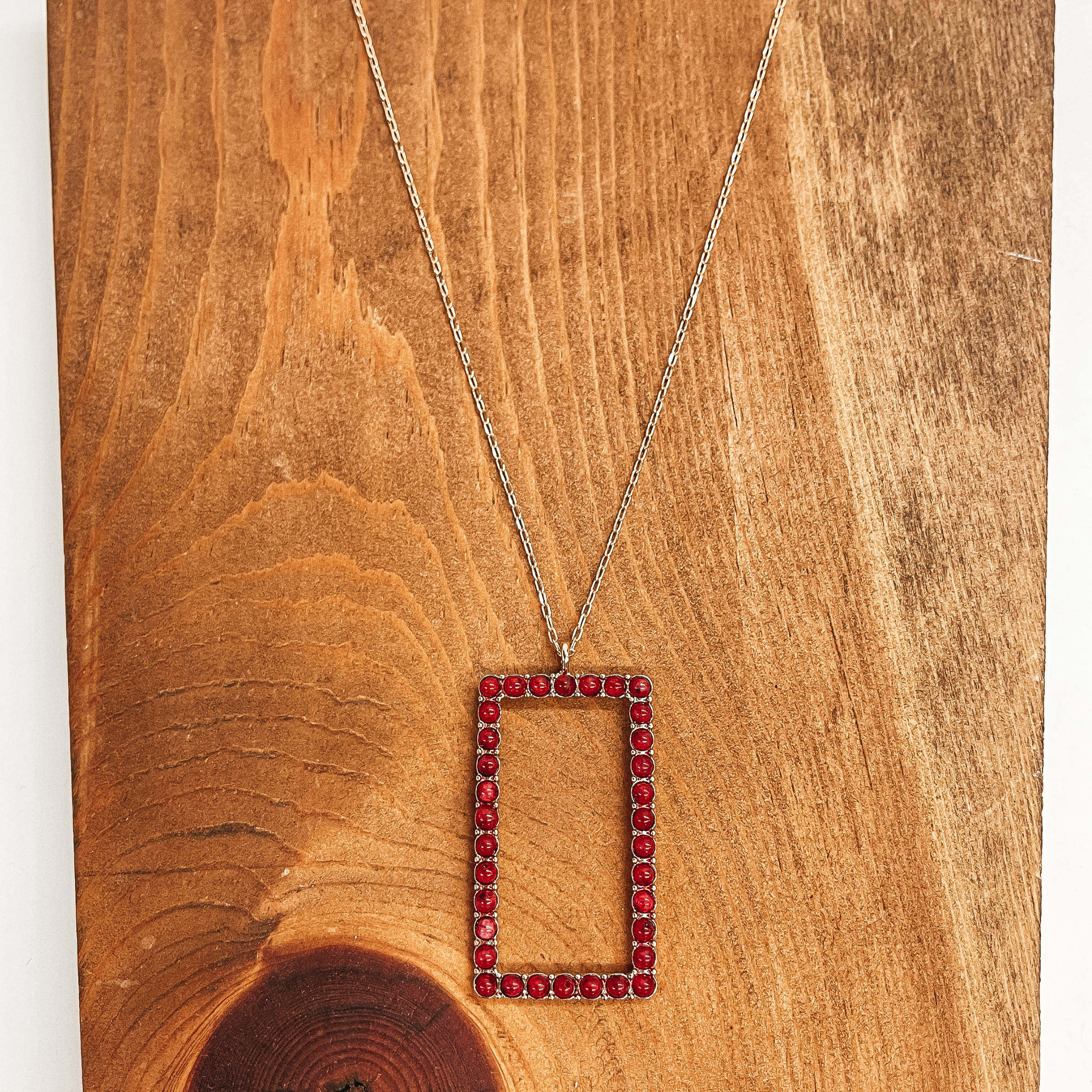 This is a long, thin, gold chain necklace with an  open rectangle pendant. The rectangle pendant has  semi-precious stones in burgundy all around. This  necklace is taken on a brown block and white  background.