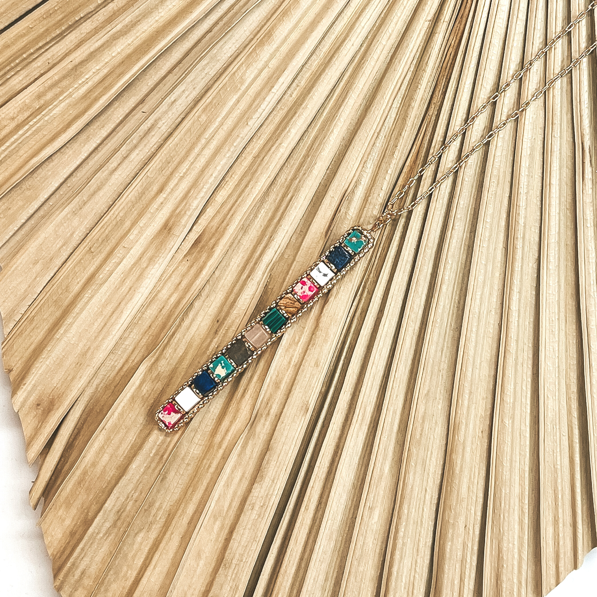 This is a long, thin, gold chain with a semi-precious bar pendant in multicolor. This necklace is  taken on a dried up palm leaf and white background.