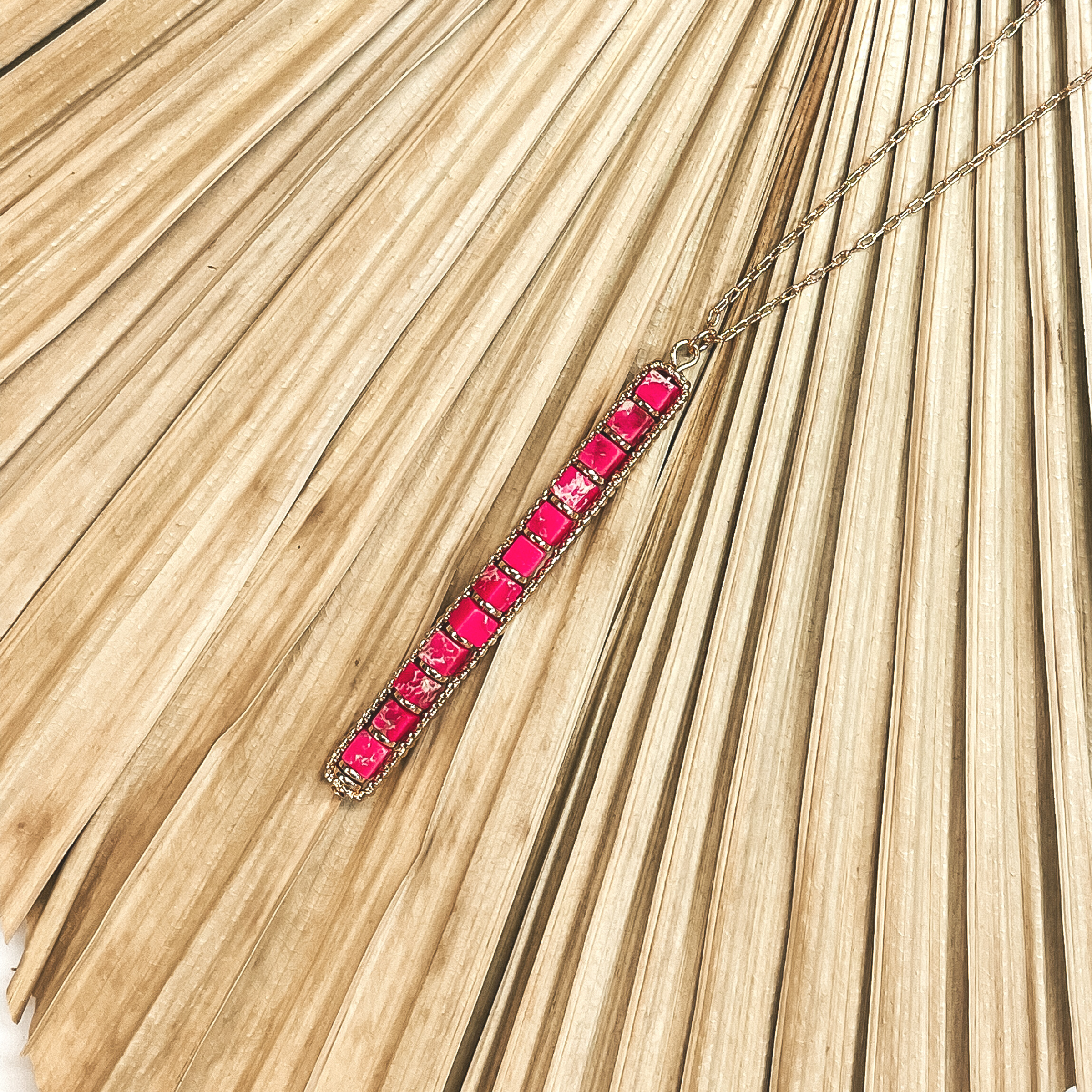 This is a long, thin, gold chain with a semi-precious bar pendant in fuchsia. This necklace is taken on a  dried up  palm leaf and white background.