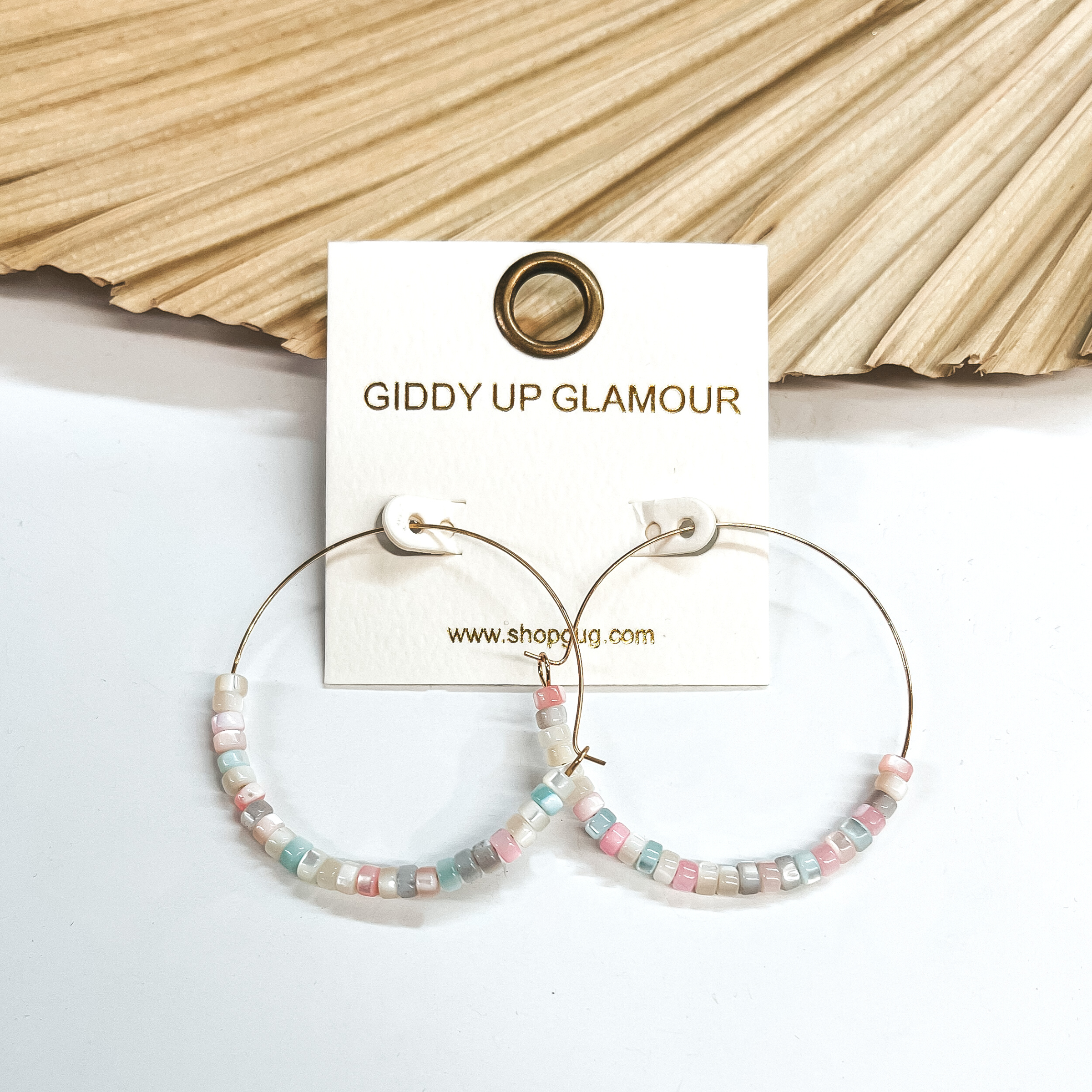 These are thin wire gold hoops with mother of pearl  beads in multicolor; ivory, light blue, light pink,  pink, and gray. The beads are in the bottom half of  the  earrings. These earrings are taken leaning up  against a dried up palm leaf and white background.