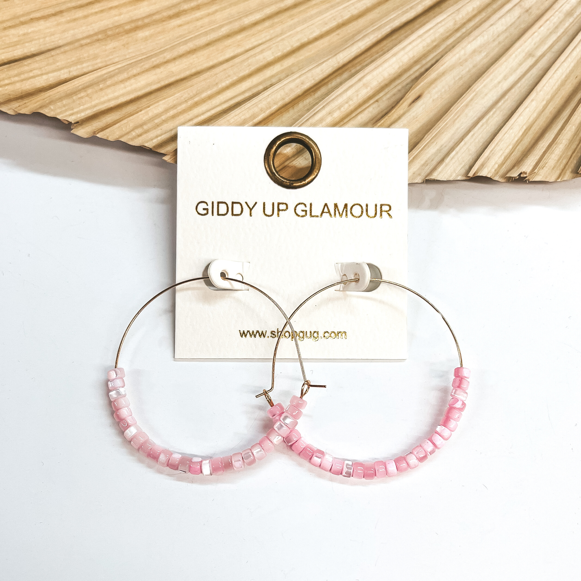 These are thin wire gold hoops with mother of pearl  beads in pink. The beads are in the bottom half of  the  earrings. These earrings are taken leaning up  against a dried up palm leaf and white background.