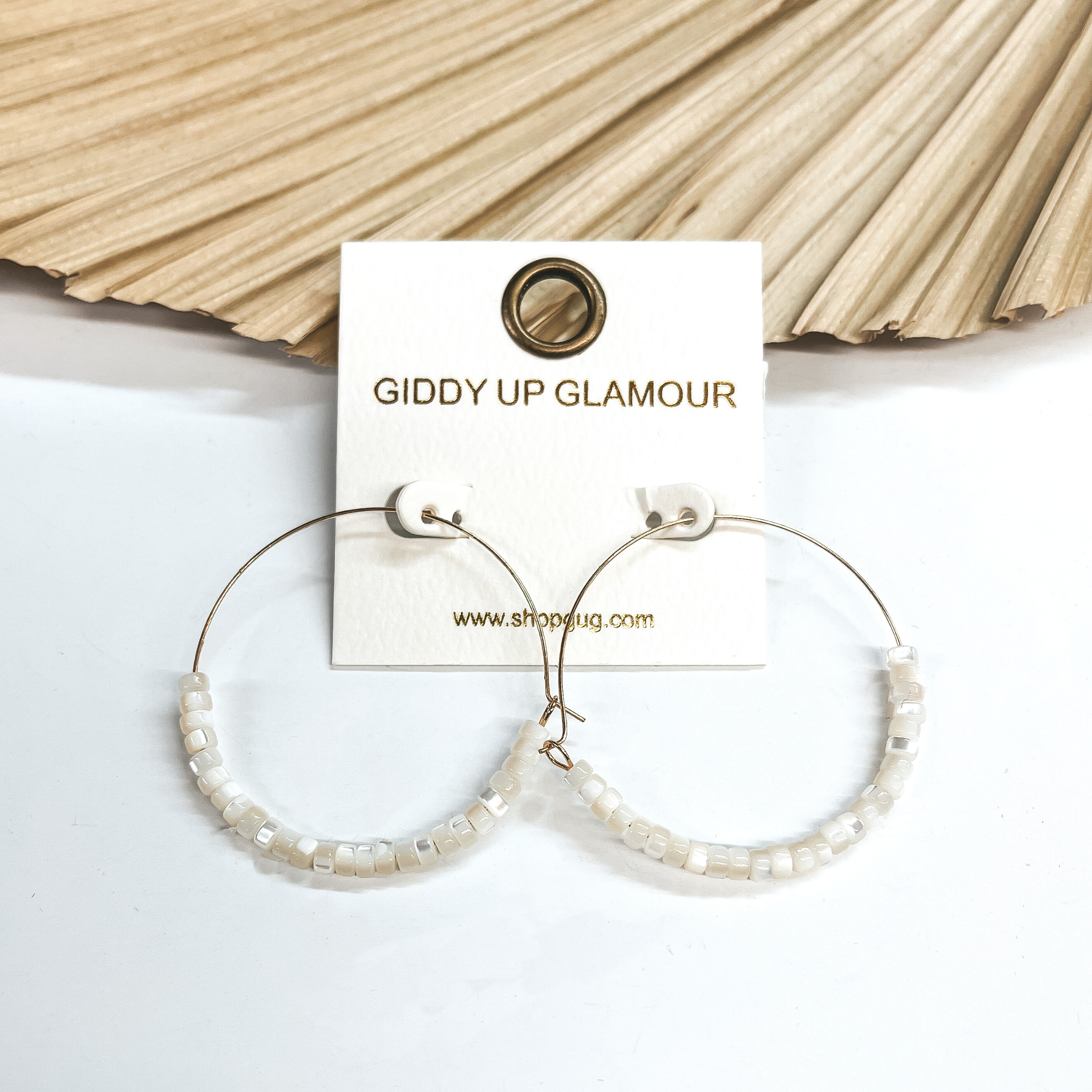 These are thin wire gold hoops with mother of pearl  beads in ivory in the bottom half of the  earrings. These earrings are taken leaning up  against a dried up palm leaf and white background.