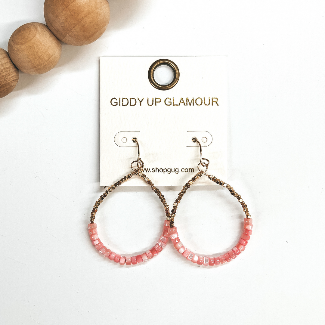 These are teardrop earrings with a fish hook backing. Bottom half has mother of pearl beads in pink  with small gold square beads in the top. These  earrings are taken on a white background and brown  wooded beads in the corner as decor.