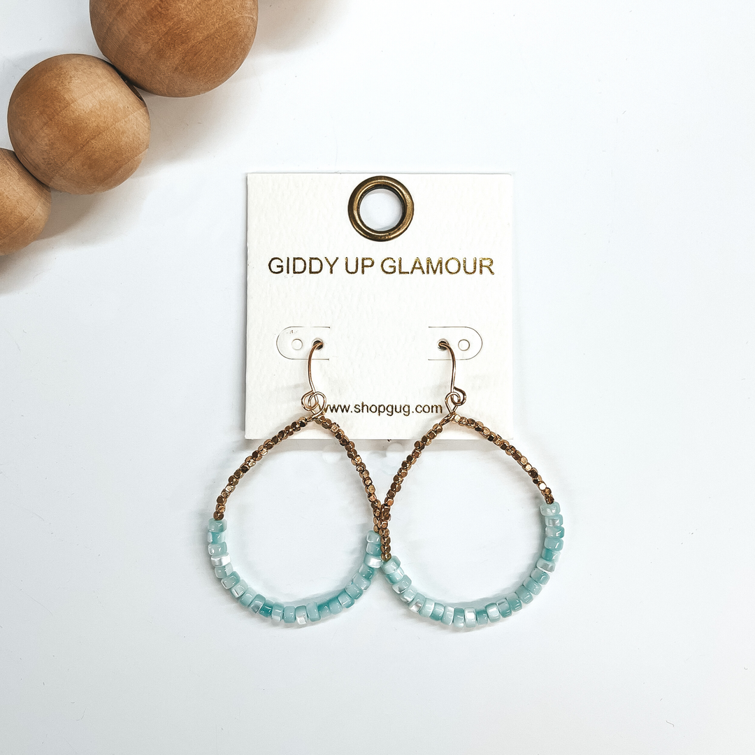 These are teardrop earrings with a fish hook backing. Bottom half has mother of pearl beads in light blue  with small gold square beads in the top. These  earrings are taken on a white background and brown  wooded beads in the corner as decor.