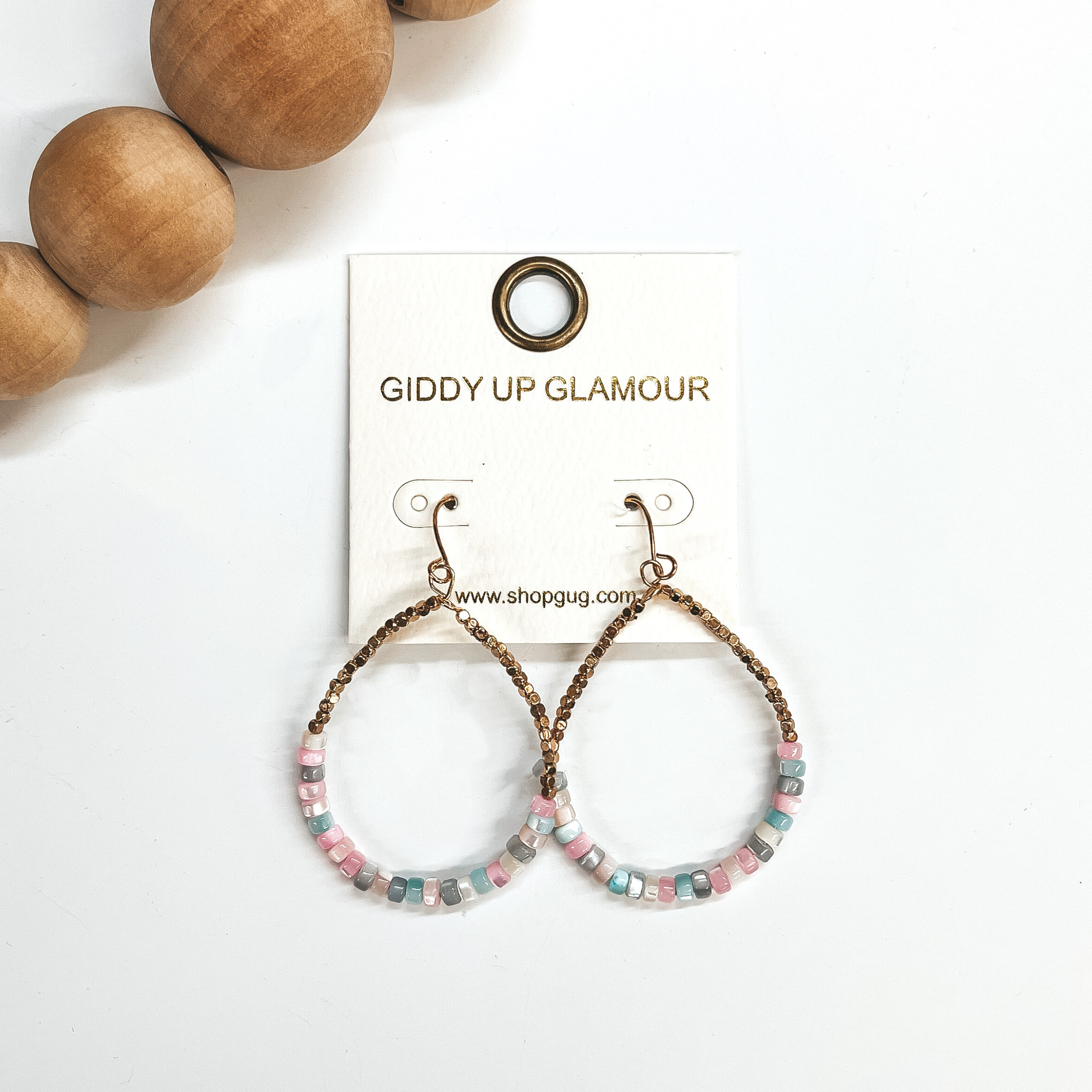 These are teardrop earrings with a fish hook backing. Bottom half has mother of pearl beads in multicolor,  light blue, light pink, pink, gray, and ivory. with small gold square beads in the top. These  earrings are taken on a white background and brown  wooded beads in the corner as decor.
