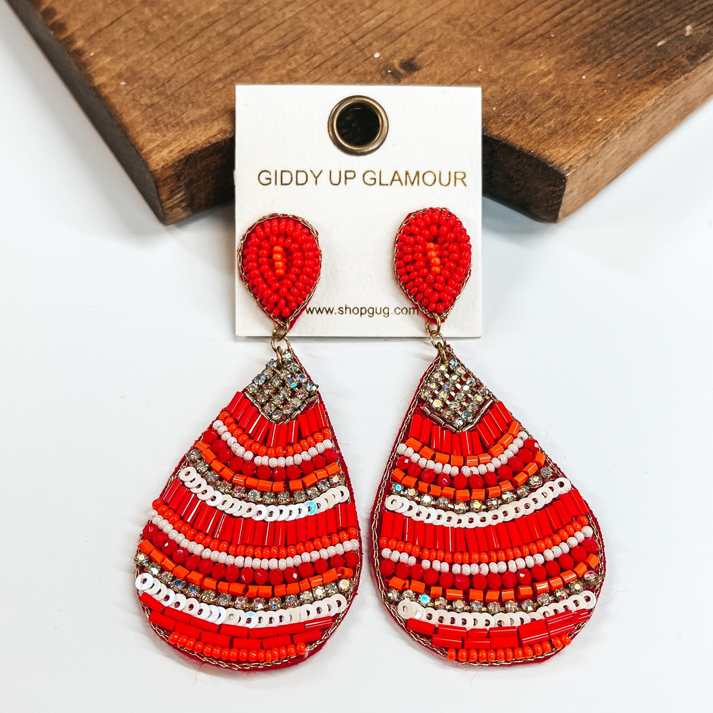 These are red teardrop earrings with a mix  of beads and crystals. There is a mix of different  sizes and colors of beads such as orange, red, and  white. There are also sequins and crystals. These  earrings are taken on a white background and leaned  up against a brown block.