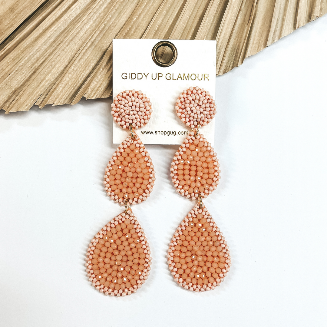 These are two tiered baby pink beaded earrings with  a post back backing. There's a mix of small matte  beads and bigger shiny beads. Taken on a white  background and leaned up against a dried up palm  leaf.