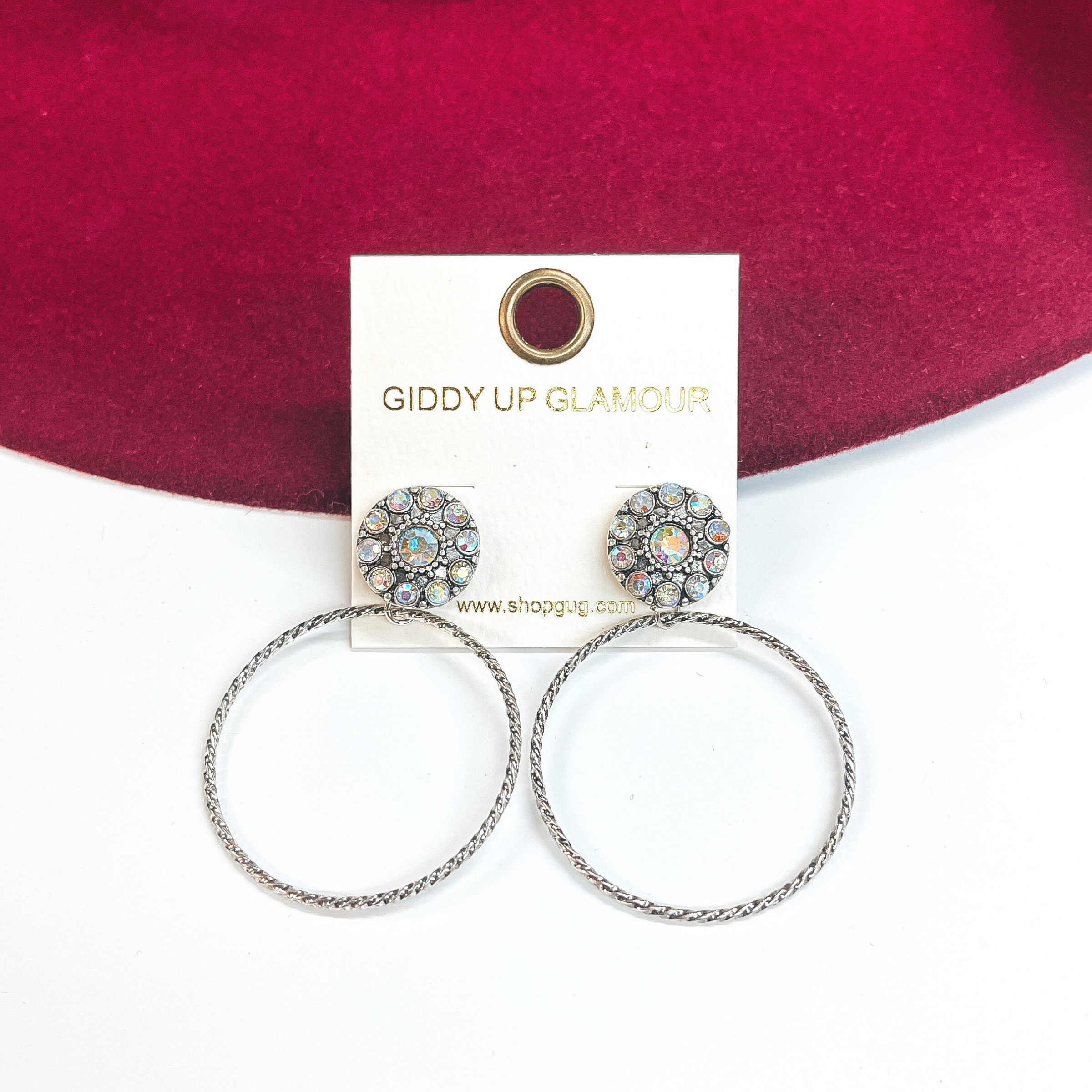 These earrings are a silver circle post back with ab  crystals, and a thin circle drop. These earrings  are taken on a white background and leaned up  against a burgundy hat brim. 