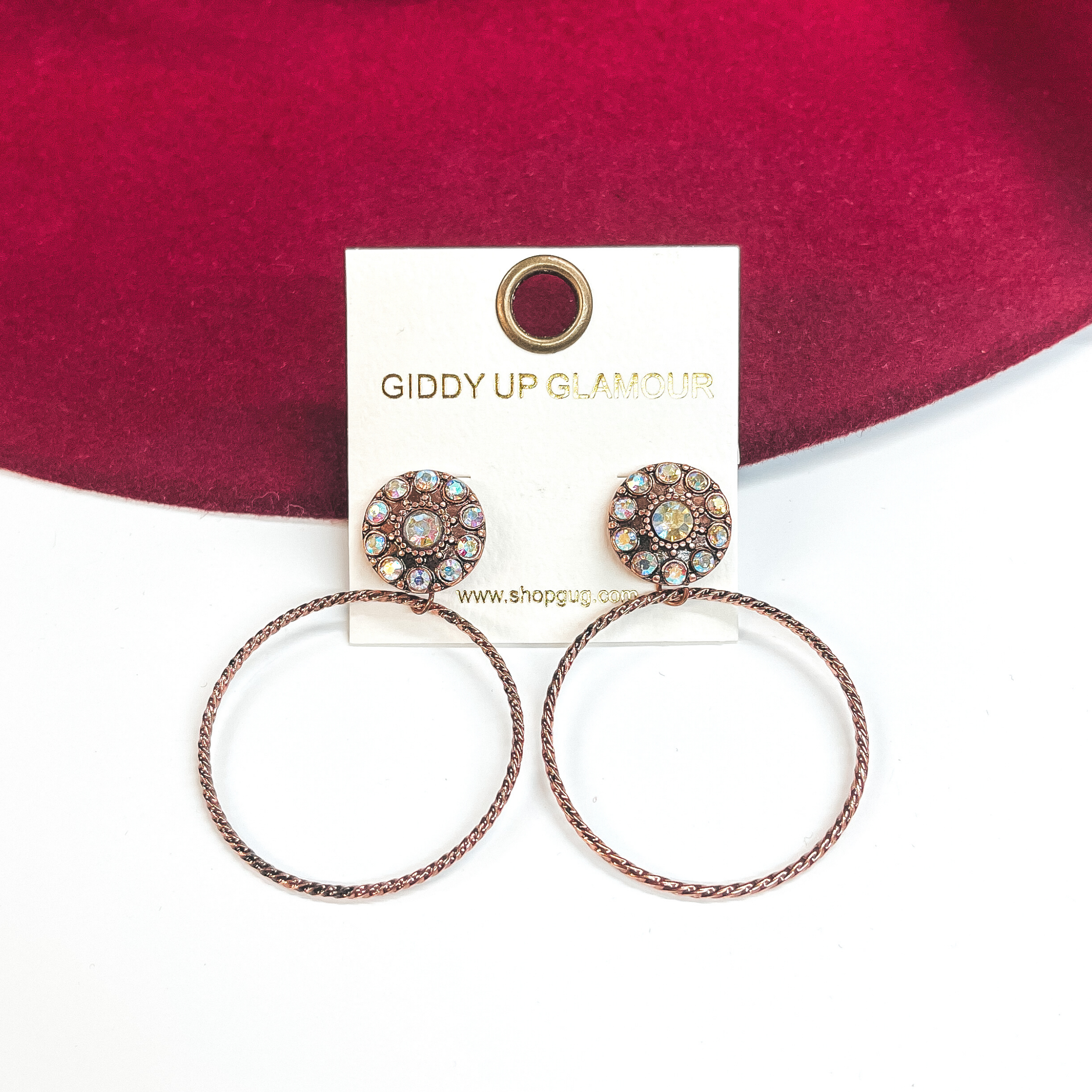 These earrings are a copper circle post back with ab  crystals, and a thin circle drop. These earrings  are taken on a white background and leaned up  against a burgundy hat brim. 