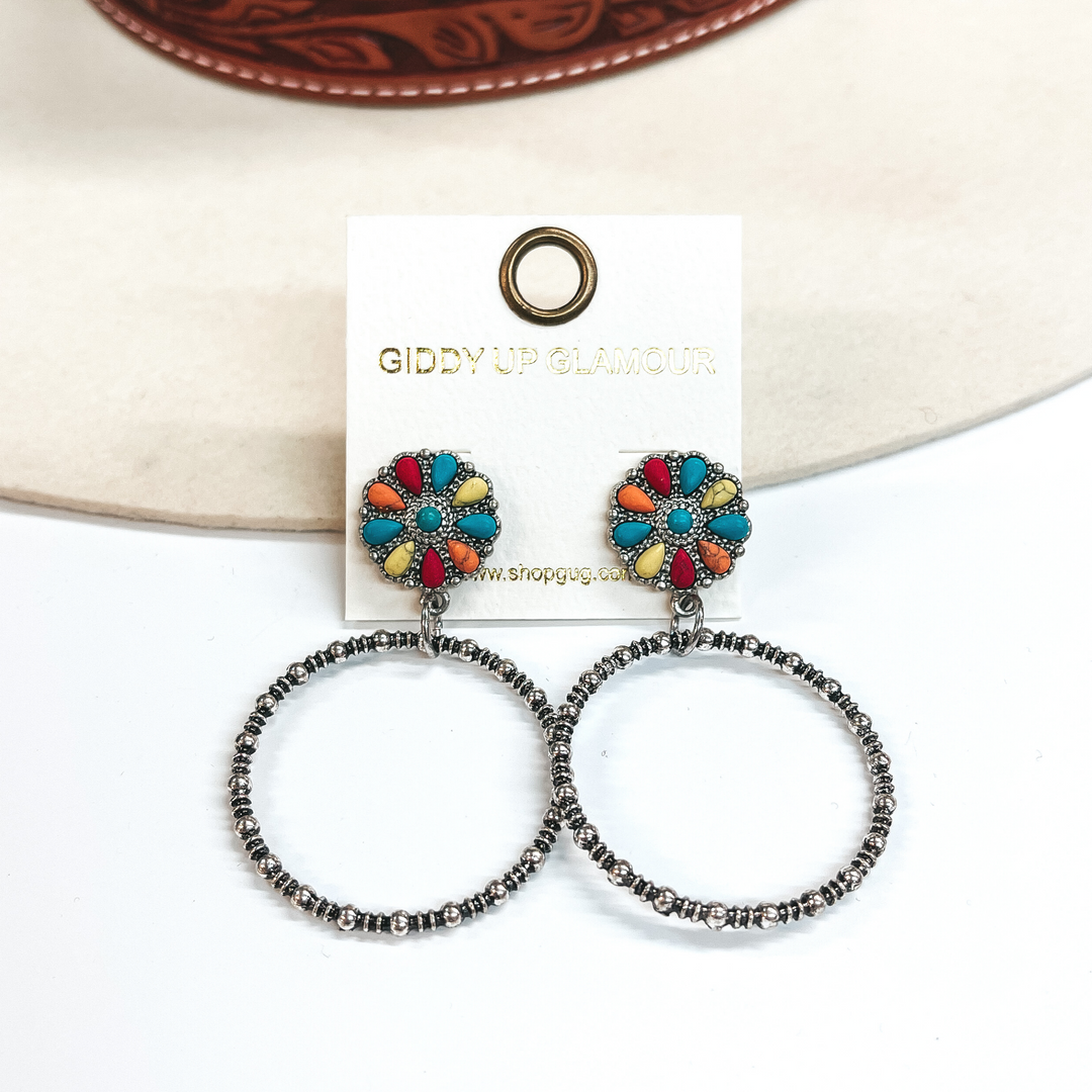 These are circle cluster post earrings with a silver  circle drop. The circle cluster has a mix of faux  colored stones in turquoise, yellow, red, and  orange. These earrings are taken on a white  background and leaned up against an ivory hat.