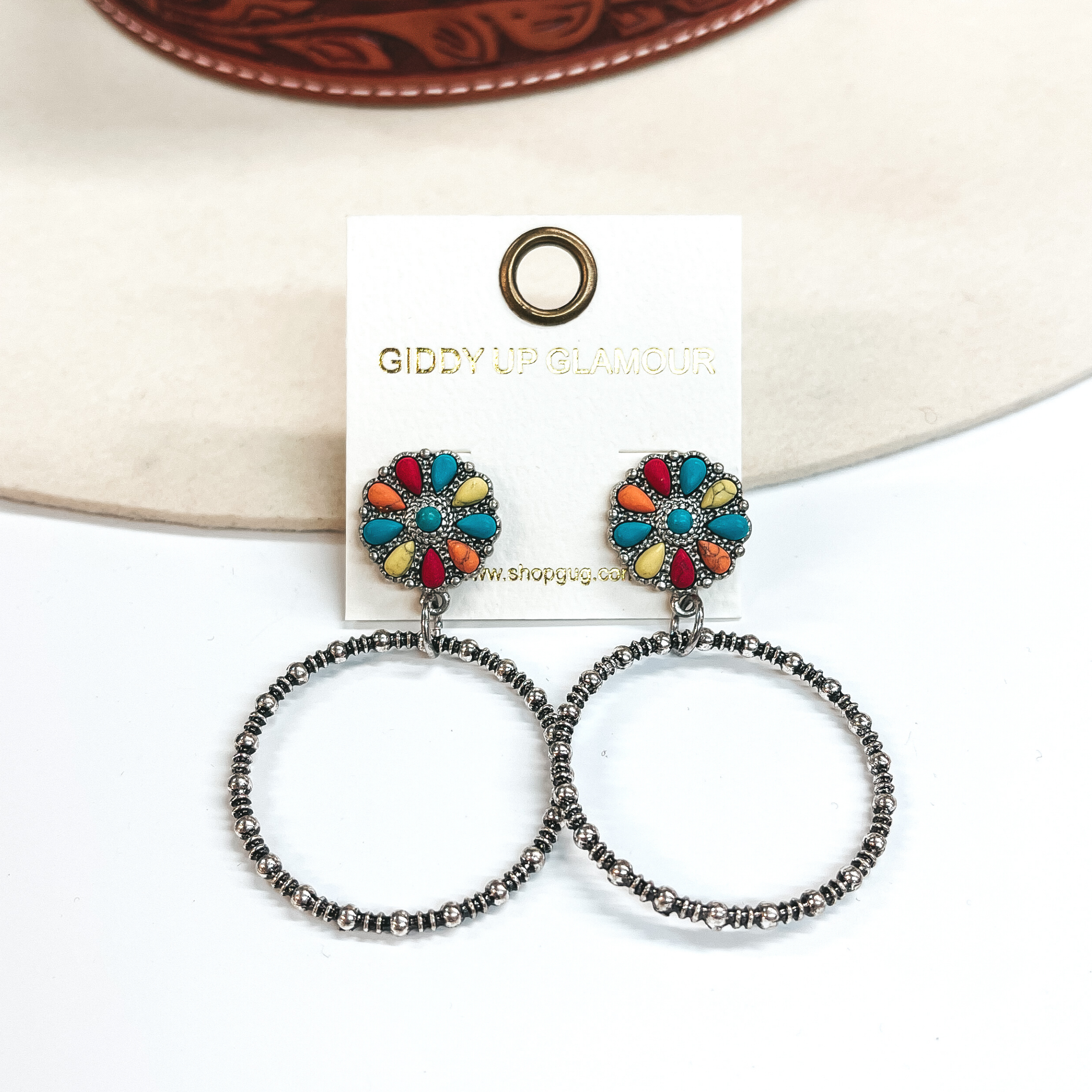These are circle cluster post earrings with a silver  circle drop. The circle cluster has a mix of faux  colored stones in turquoise, yellow, red, and  orange. These earrings are taken on a white  background and leaned up against an ivory hat.