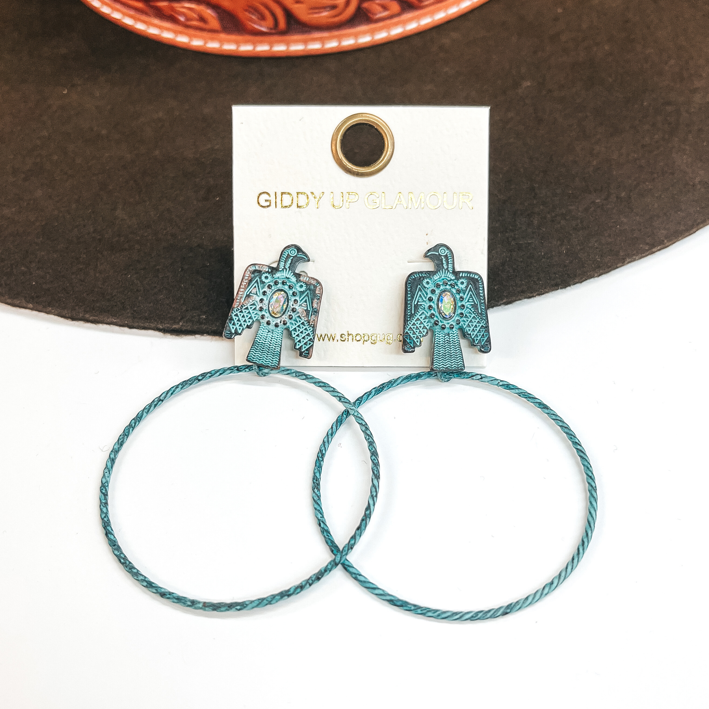 These are thunderbird post earrings with an AB  crystal in the center and a thin  circle drop in patina. These earrings are taken on  a white background and leaned up against a dark  brown hat. 