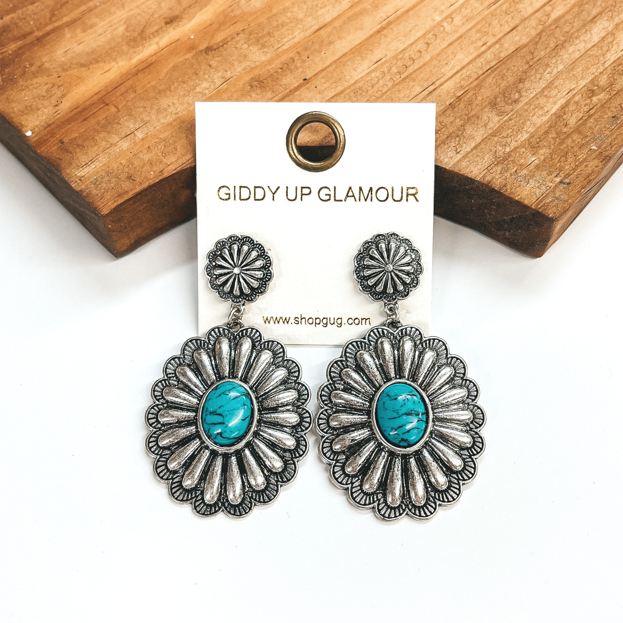 These are silver concho post earrings with a large  concho drop with a faux turquoise stone in the  center. These earrings are taken on a white  background and leaned up against a brown block.