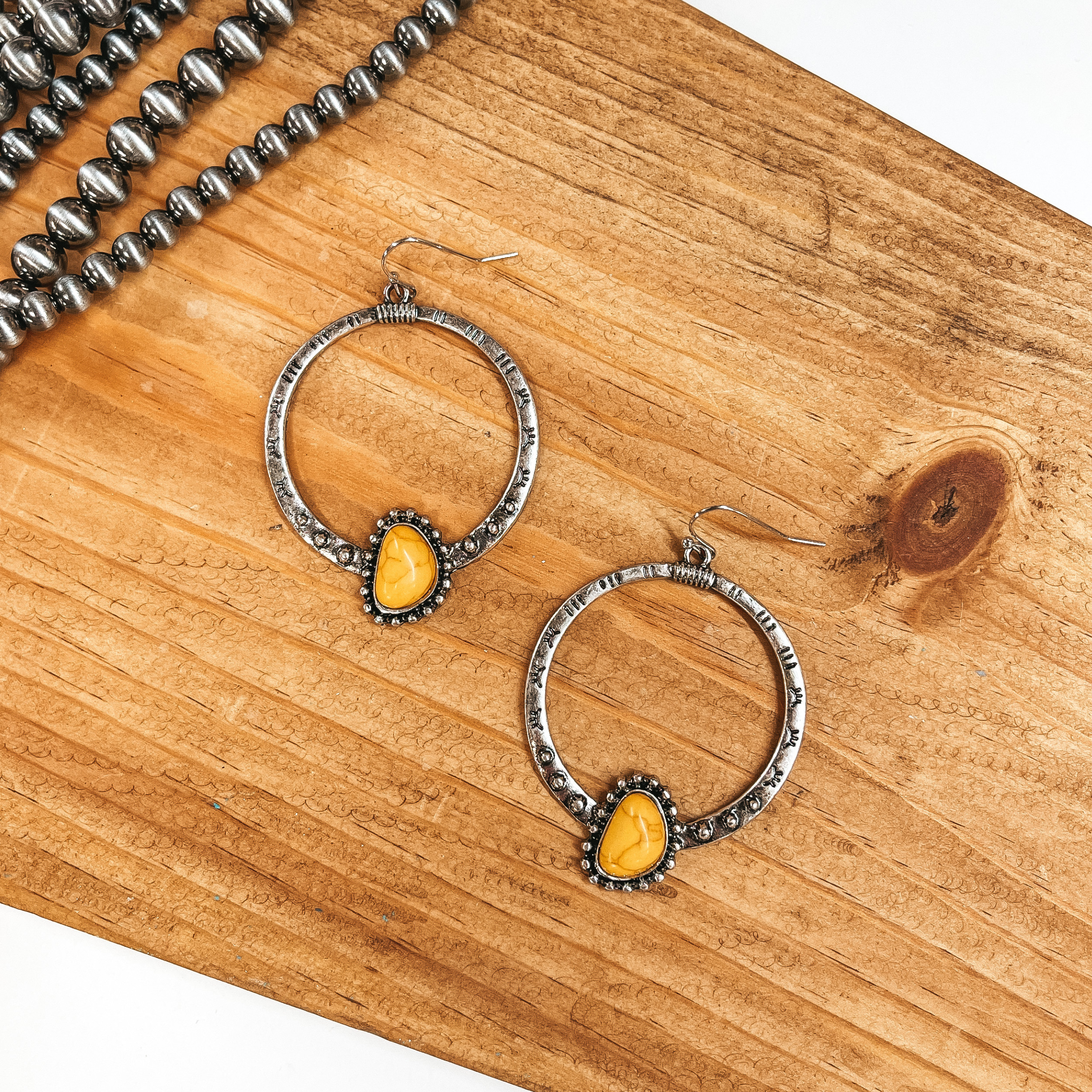 These are silver fish hook earrings with a circle  drop. The hoop has western engravings and a semi  circle stone in mustard yellow with silver  details around. These earrings are taken on a brown  block and faux navajo pearls as decor in the side.