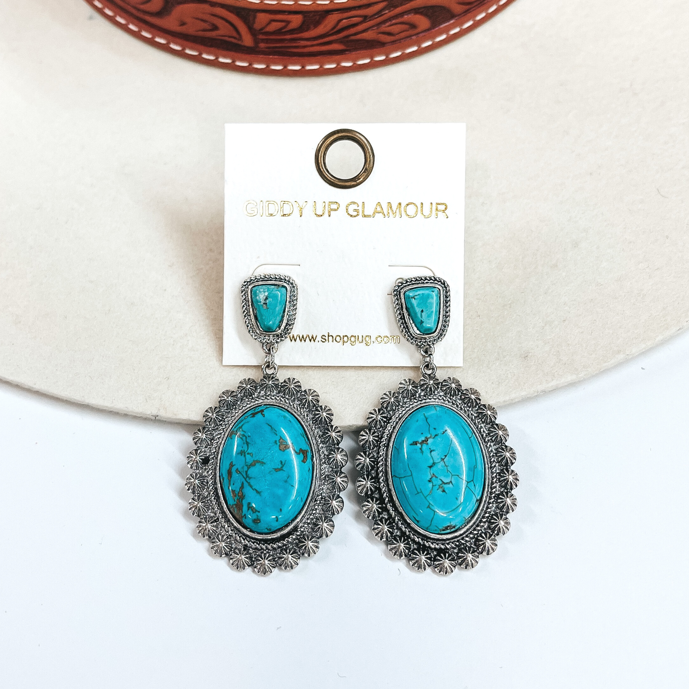 These are western silver post earrings with  faux turquoise stones. It has a small stone in a  silver textured setting and an oval drop, the oval  drop has a large oval turquoise stone in a silver western textured setting. These earrings are taken  on an ivory felt hat and a white background.