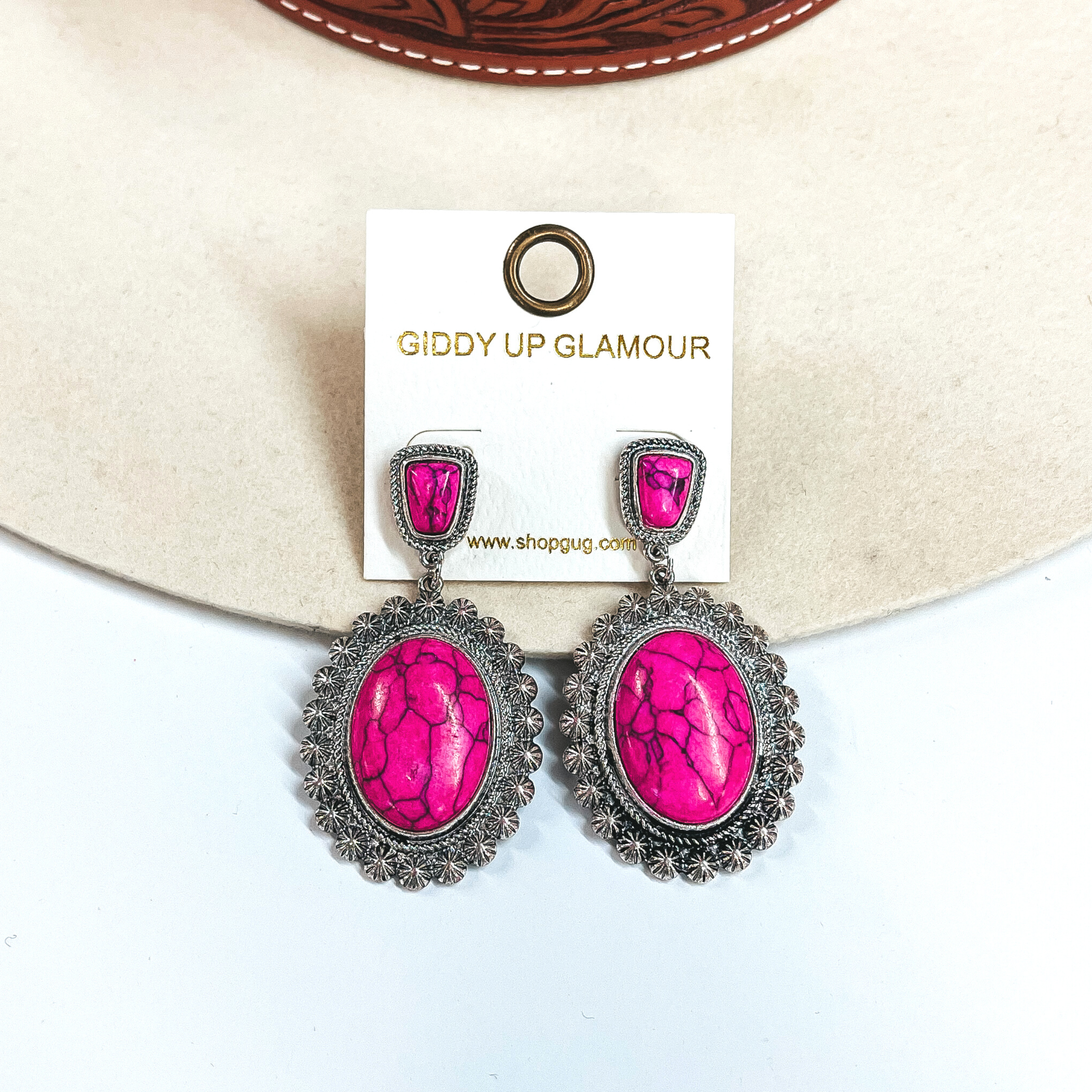 These are western silver post earrings with  faux pink stones. It has a small stone in a  silver textured setting and an oval drop, the oval  drop has a large oval pink stone in a silver western textured setting. These earrings are taken  on an ivory felt hat and a white background.