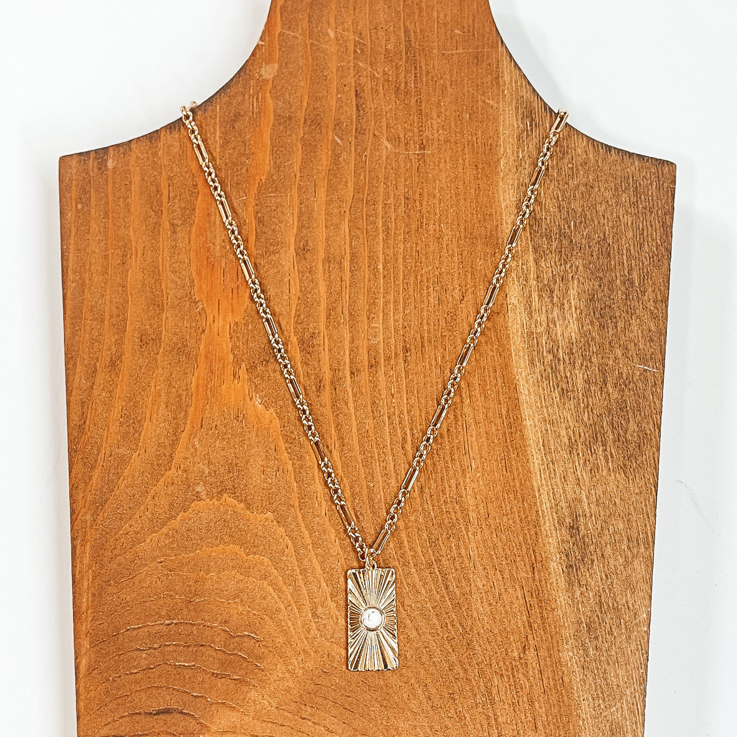This is a gold chain necklace with a rectangle  pendant, the pendant has a sunburst texture and  a small stone in white. This necklace is taken on  a brown necklace board and a white background.
