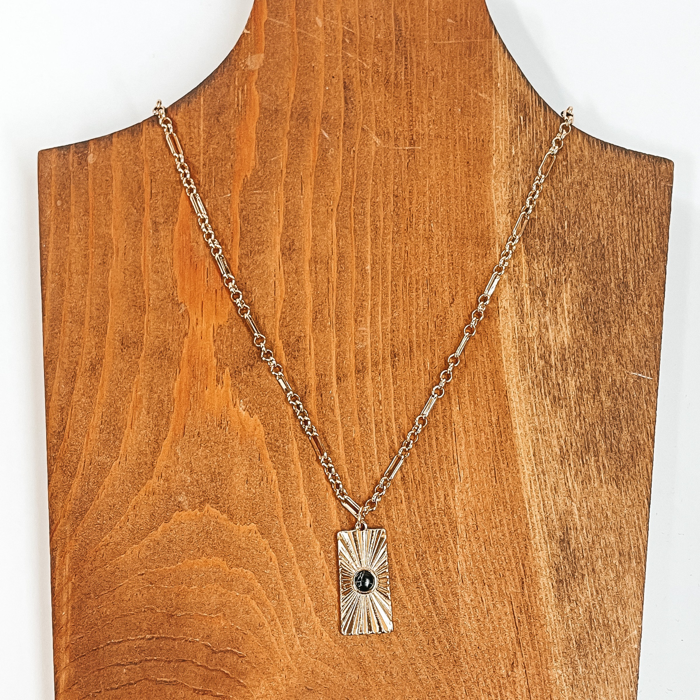 This is a gold chain necklace with a rectangle  pendant, the pendant has a sunburst texture and  a small stone in black. This necklace is  taken on  a brown necklace board and a white background.