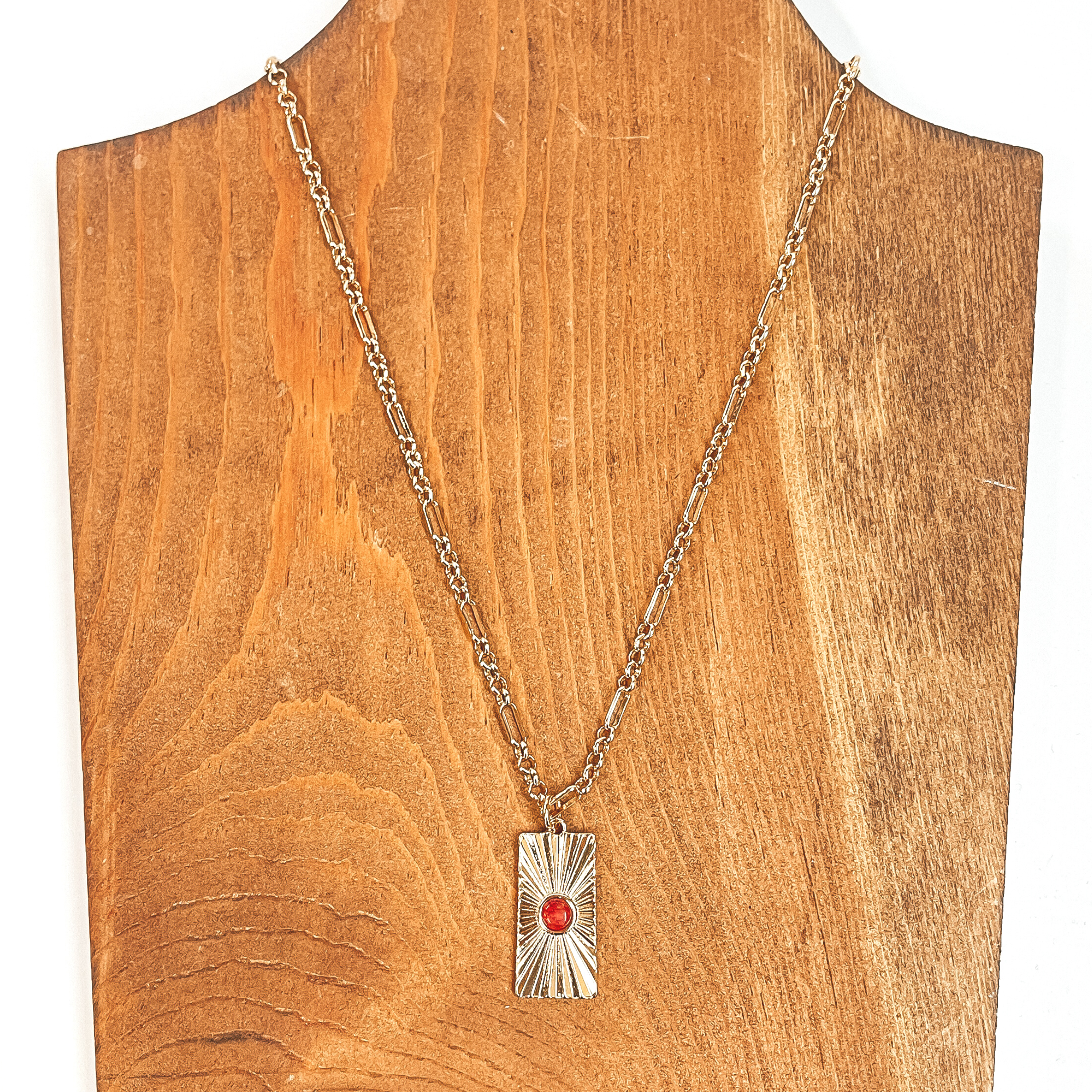 This is a gold chain necklace with a rectangle  pendant, the pendant has a sunburst texture and  a small stone in red. This necklace is taken on  a brown necklace board and a white background.