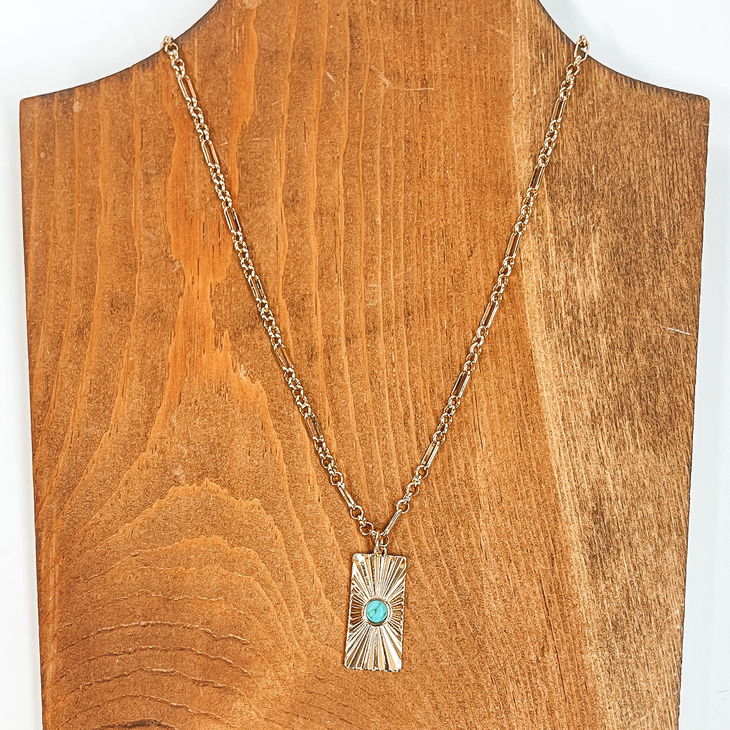 This is a gold chain necklace with a rectangle  pendant, the pendant has a sunburst texture and  a small stone in turquoise. This necklace is  taken on  a brown necklace board and a white background.