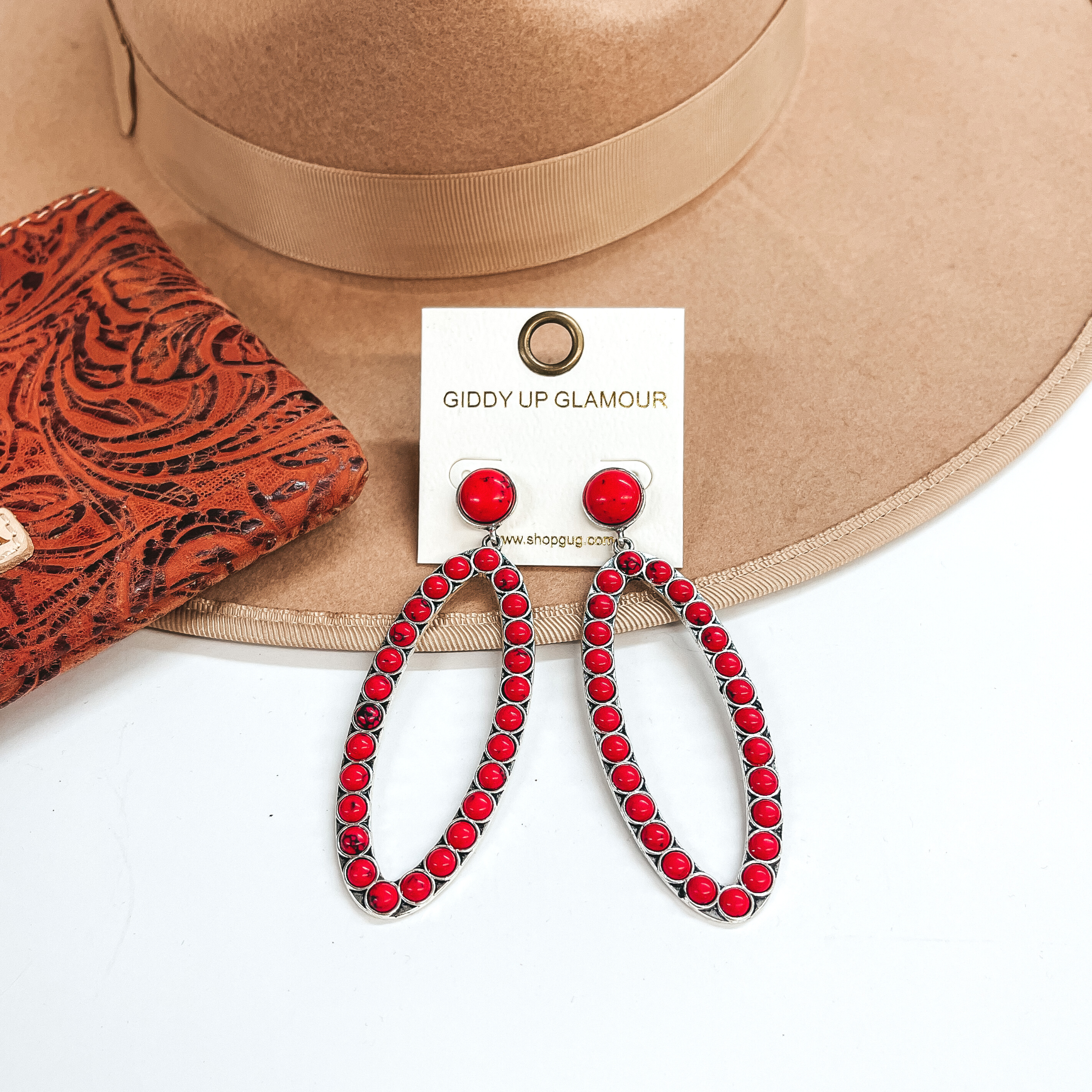 These are open oval drop earrings with red stones.  Post back earrings with a small stone and an open  oval drop with red stones all around. These are  taken on a light brown hat brim and white background.  There is a leather printed wallet in the side  as decor.