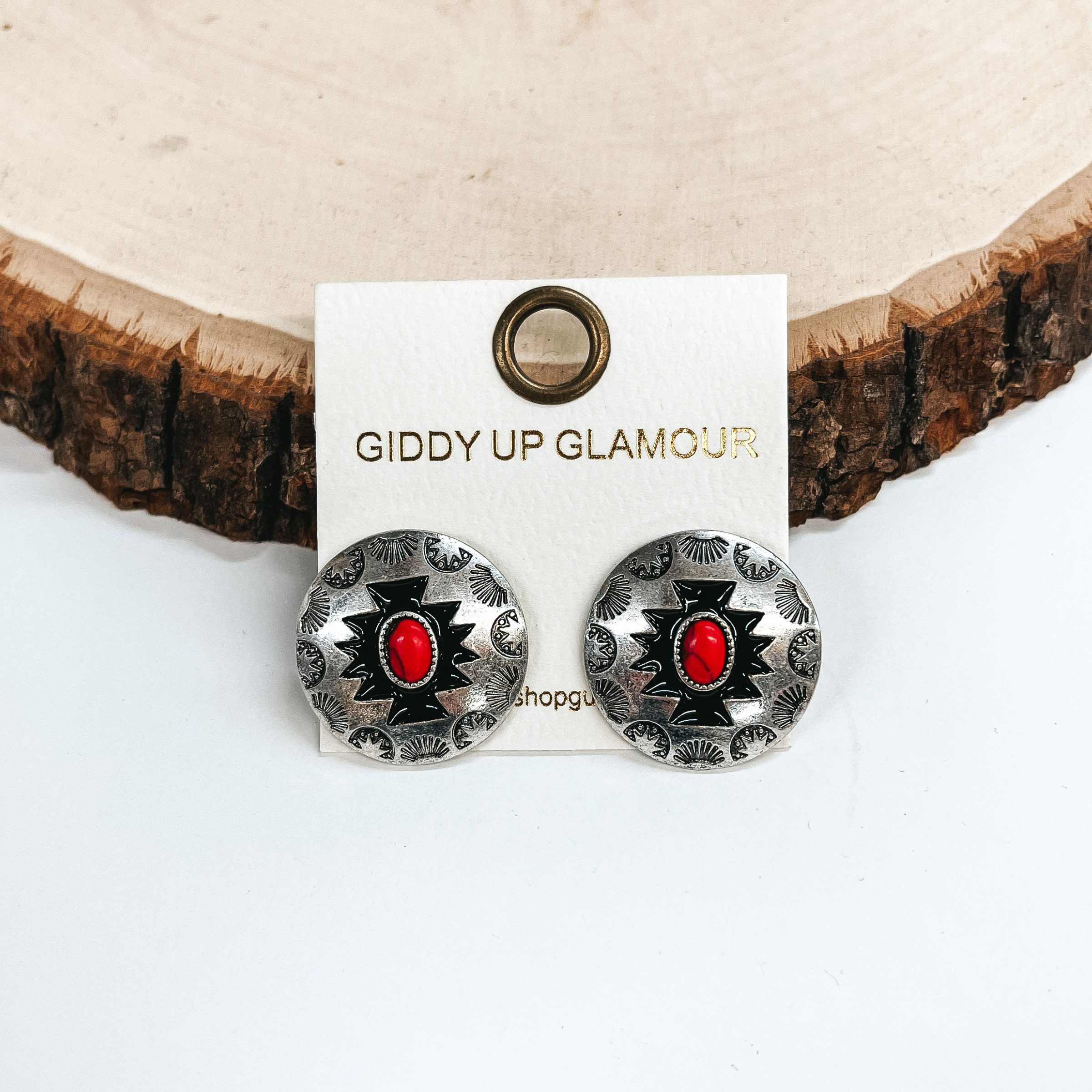 These are silver circle post back earrings with  aztec design and a stone in the center, the stone  is red. These earrings are taken on a white  background and leaned up against a slab of wood.