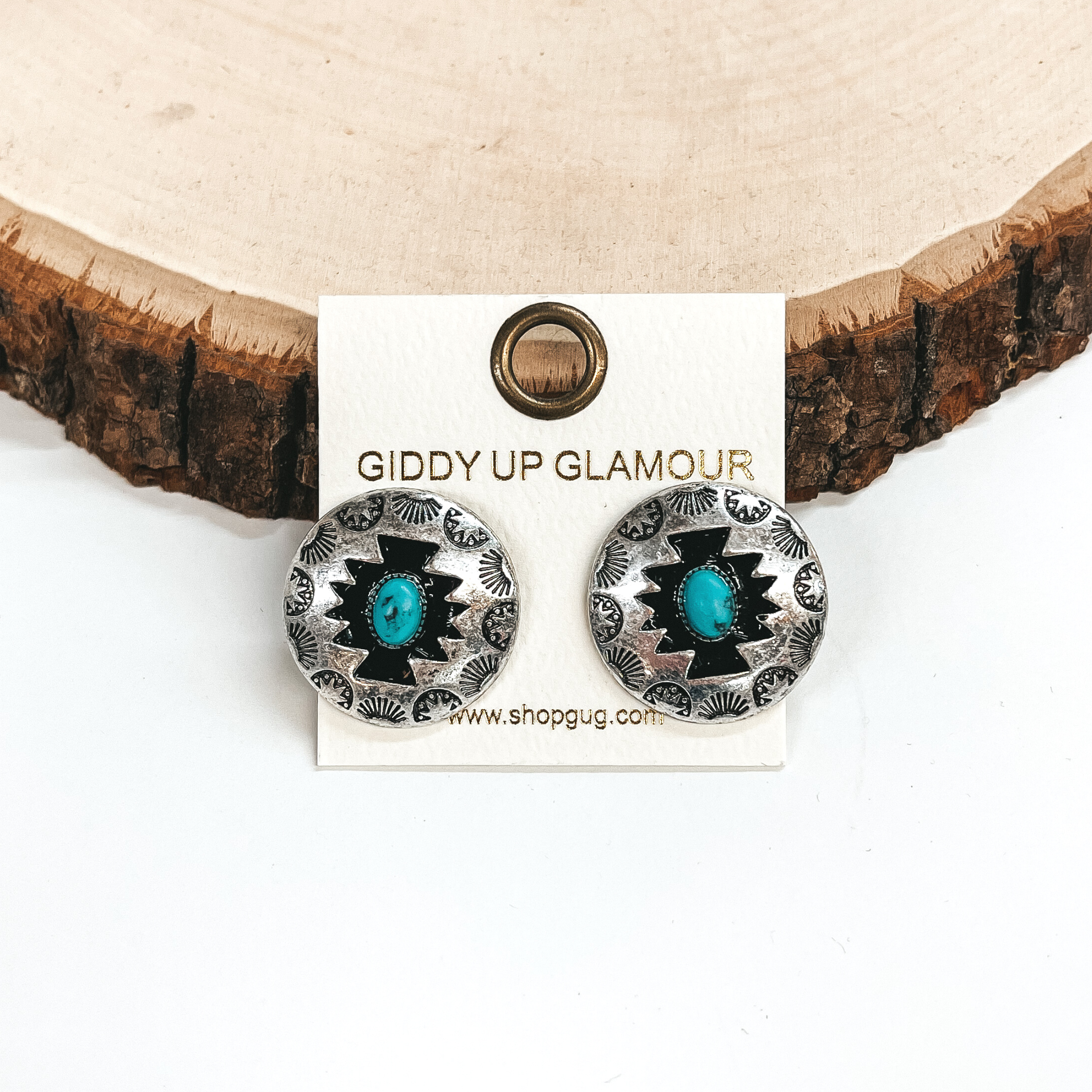 These are silver circle post back earrings with  aztec design and a stone in the center, the stone  is turquoise. These earrings are taken on a white  background and leaned up against a slab of wood.