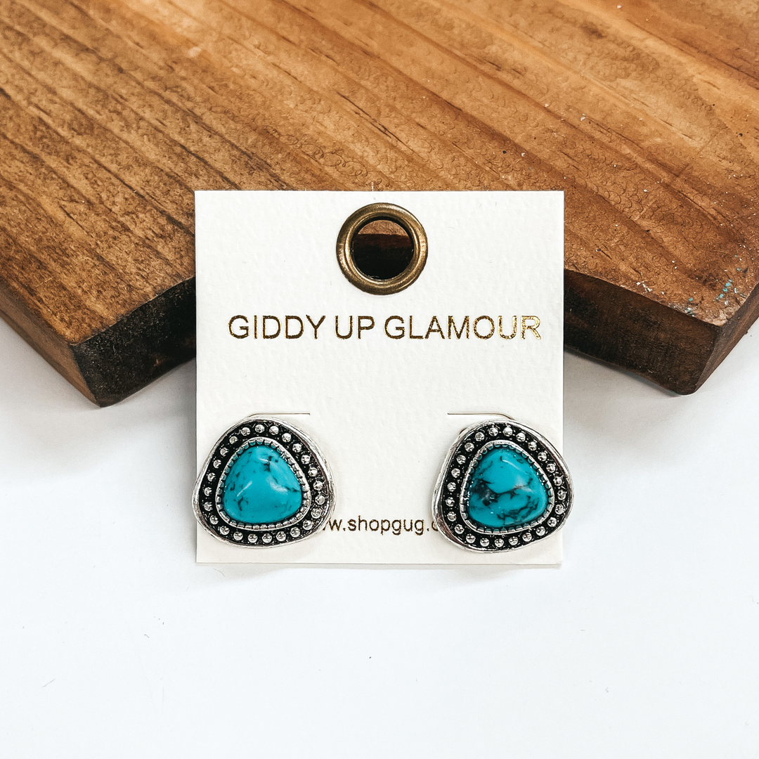 These are triangular post back silver earrings  with detailing and faux stone in turquoise. These  earrings are taken on a white background and  leaned up against a brown block.
