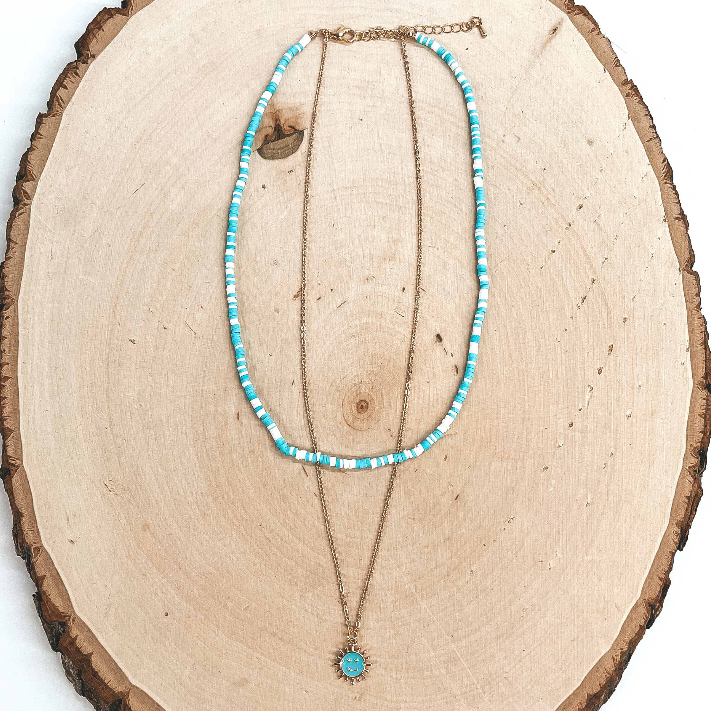 This is a double layered necklace the smaller  necklace has rubber beads in white and turquoise.  The bigger necklace is a small gold chain with a  turquoise sun pendant with a happy face. This  necklace is taken on a slab of wood and a white  background.