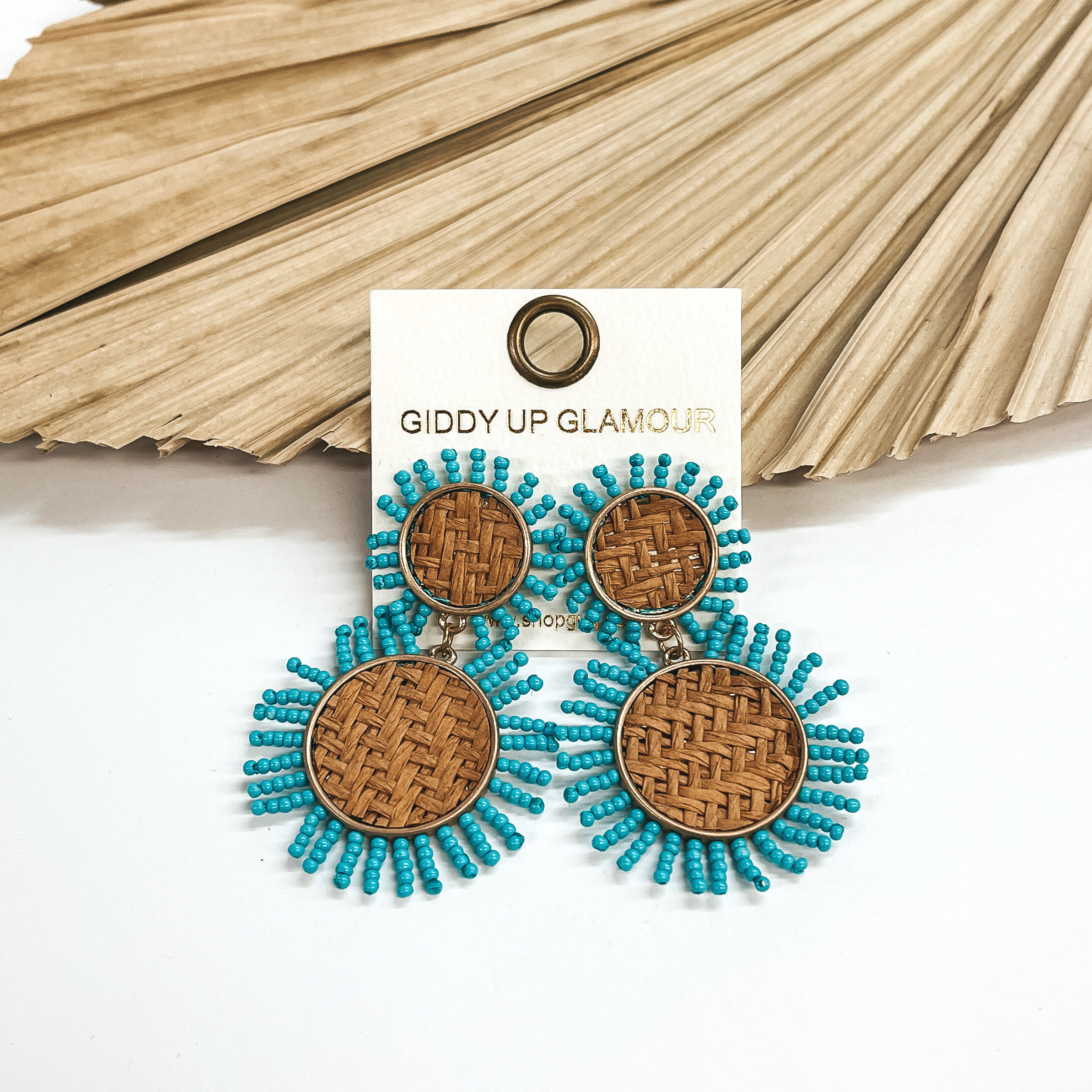 These earrings are two tiered woven earrings in a  gold setting with turquoise seed beads all around.  These earrings are taken on a white background and  leaned up against a dried up palm leaf.