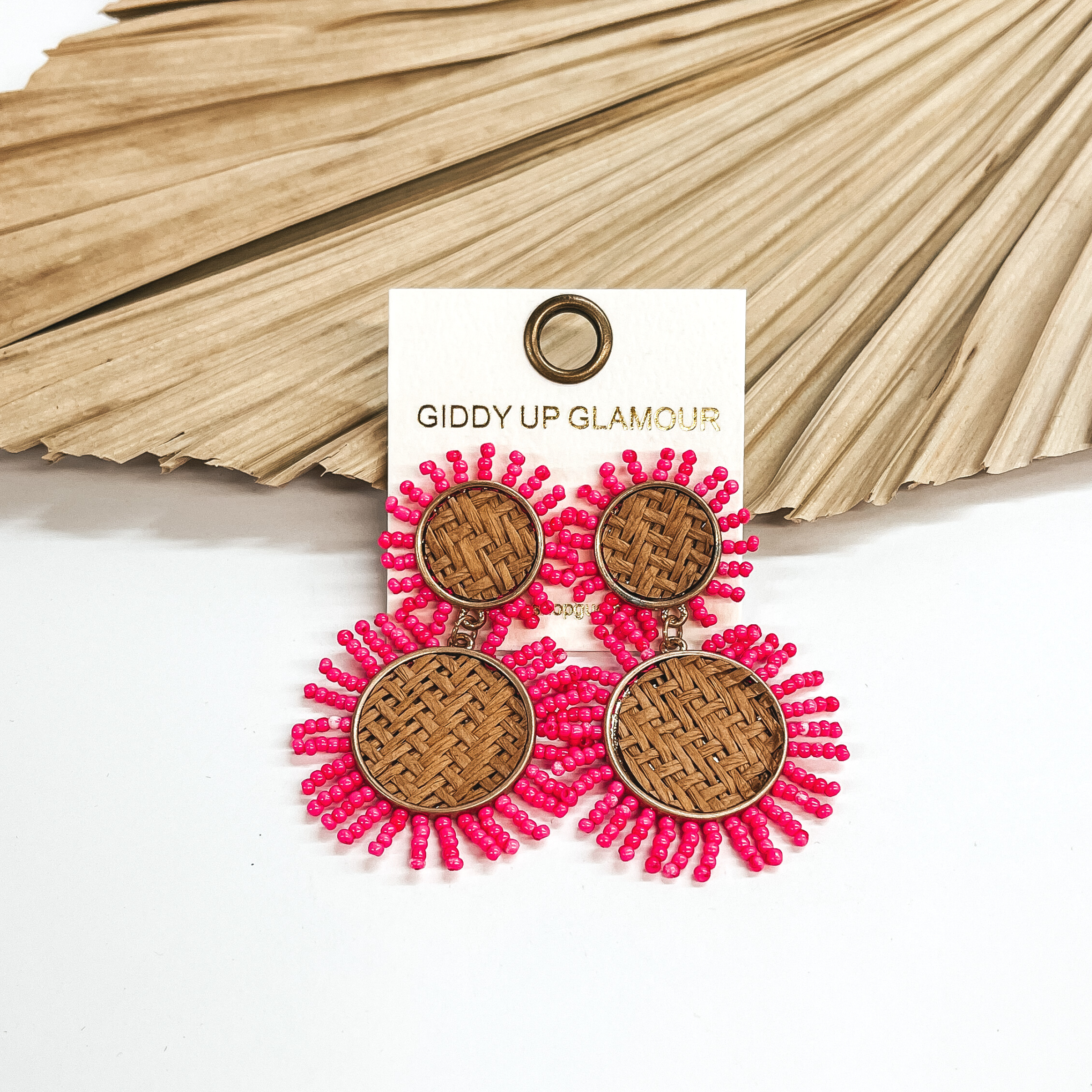 These earrings are two tiered woven earrings in a  gold setting with pink seed beads all around. These  earrings are taken on a white background and leaned  up against a dried up palm leaf.
